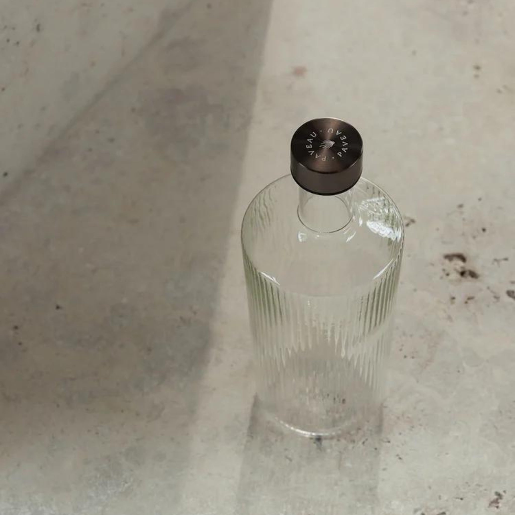 De Paveau Waterfles White Haven 1,25 l van Paveau, met verticale ribbels en een donkere ronde kap, staat op een licht gestructureerd oppervlak met vage vlekken en schaduwen.