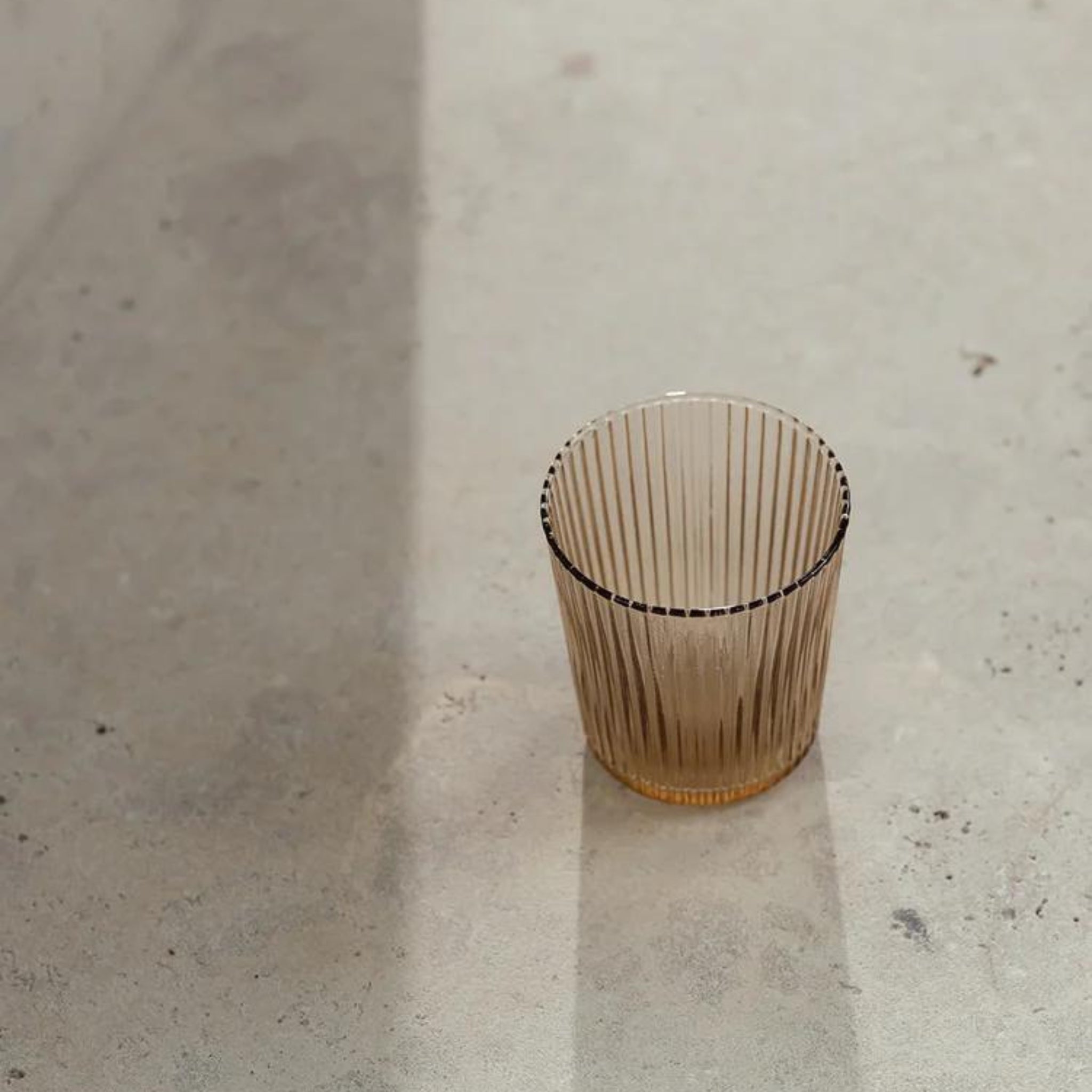 De Paveau Waterglas Bells Beige van Paveau, een transparant, geribbeld glas met een amberkleurige tint, rust op een zonovergoten betonnen oppervlak en werpt een zachte schaduw.