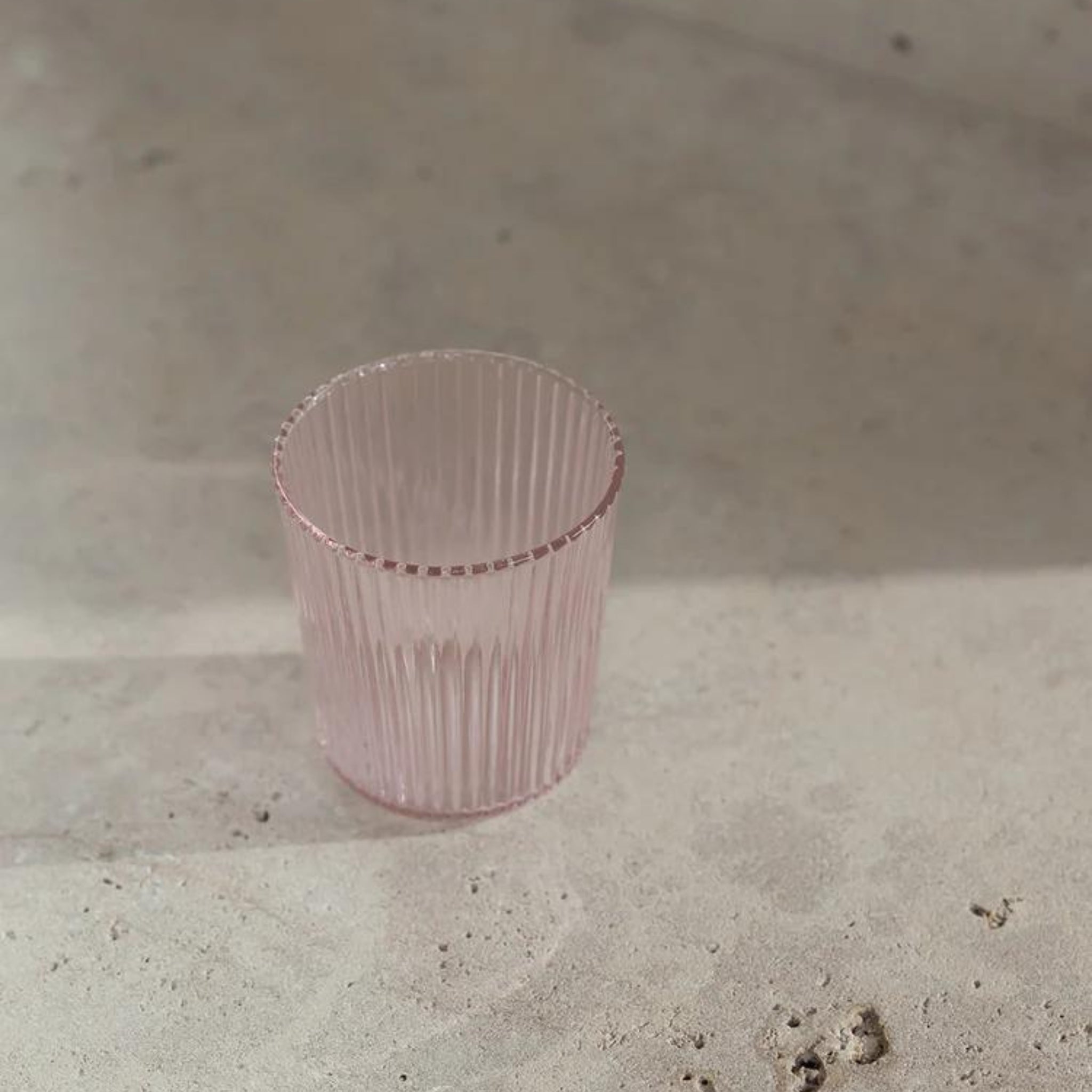 De Paveau Waterglas Pink van Paveau, een lichtroze geribbelde glazen beker, staat leeg op een licht gestructureerd oppervlak in zacht natuurlijk licht, en werpt een subtiele schaduw.