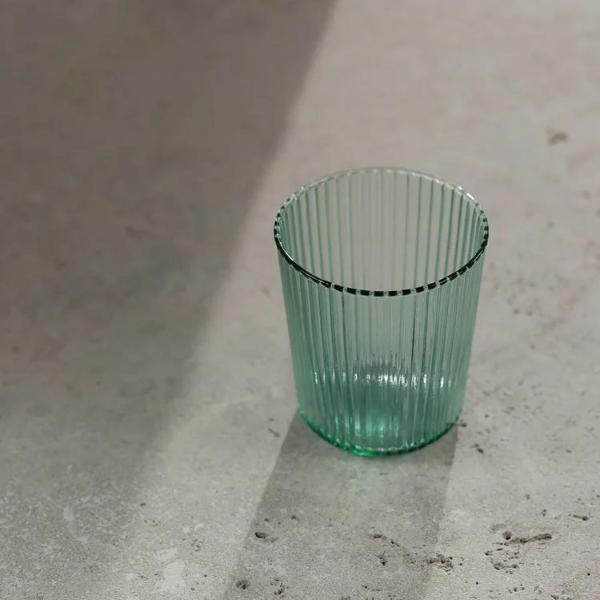 Een Paveau Waterglas Bondi Green tumbler van Paveau staat op een gestructureerd, licht oppervlak, gedeeltelijk in de zon met zijn schaduw geworpen naast andere duurzame glazen. Het geribbelde groen getinte glas voegt een frisse toets toe aan het tafereel.