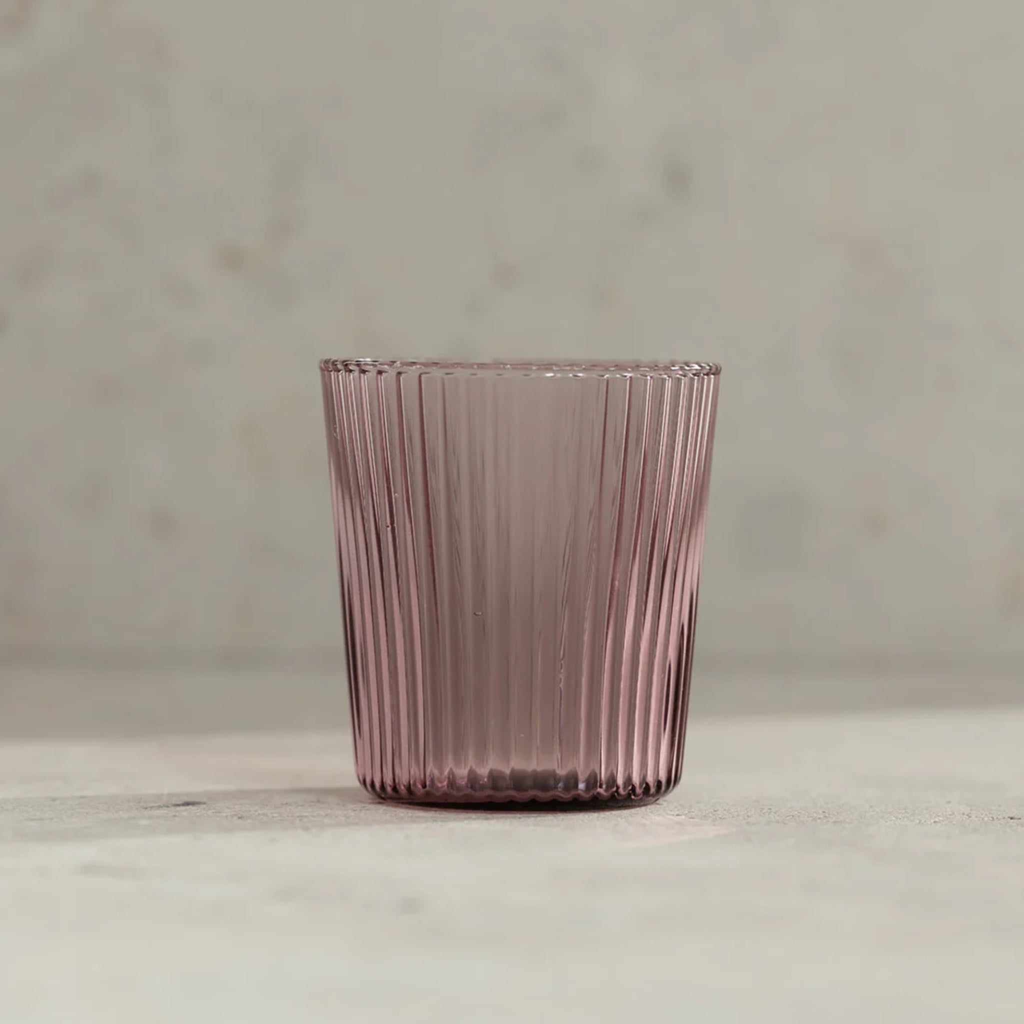 De Paveau Waterglas Broome van Paveau, met een roze tint en verticaal geribbeld ontwerp, staat op een zacht neutraal oppervlak - ideaal voor iedereen die op zoek is naar een stijlvolle, duurzame drinkbril.