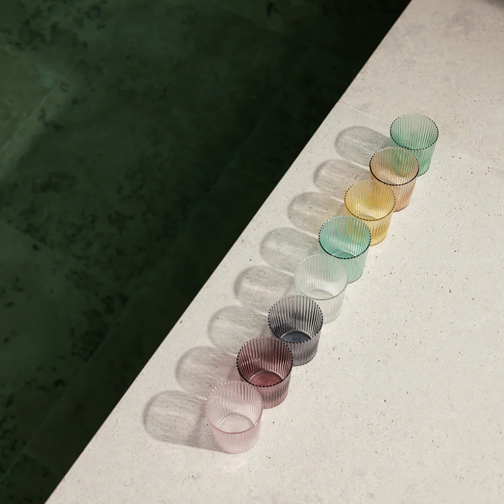 Acht geribbelde, doorschijnende Paveau Waterglas Broome by Paveau in verschillende kleuren staan diagonaal opgesteld op een licht oppervlak met zachte schaduwen, naast een donkergroene tegelvloer.