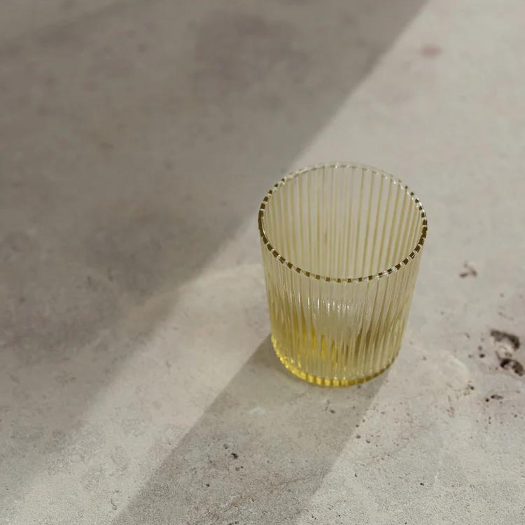 De Paveau Waterglas Cable Yellow, een transparant geribbeld geel glas van Paveau, ligt op een licht oppervlak en werpt een zachte schaduw in het zonlicht. Ontdek de duurzame Paveau waterglas voor elke gelegenheid.