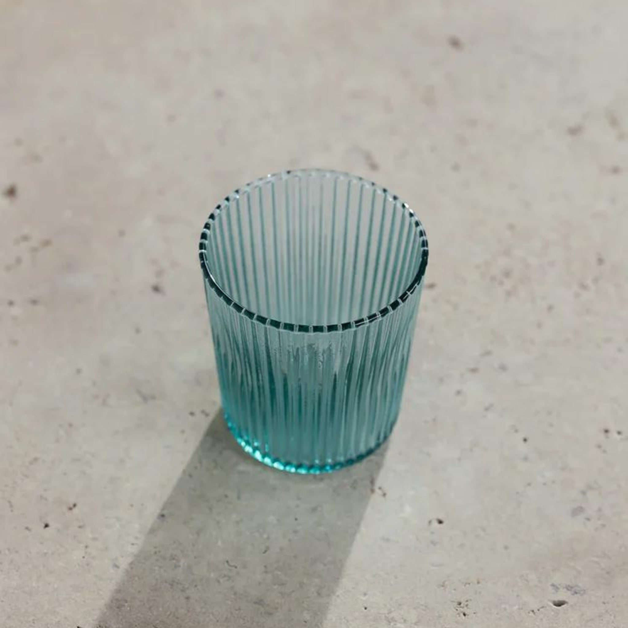 De Paveau Waterglas Lucky Turquoise van Paveau rust op een lichtbeige gestructureerd oppervlak. Het transparante, geribbelde groenblauwe glas werpt een zachte schaduw naar links.