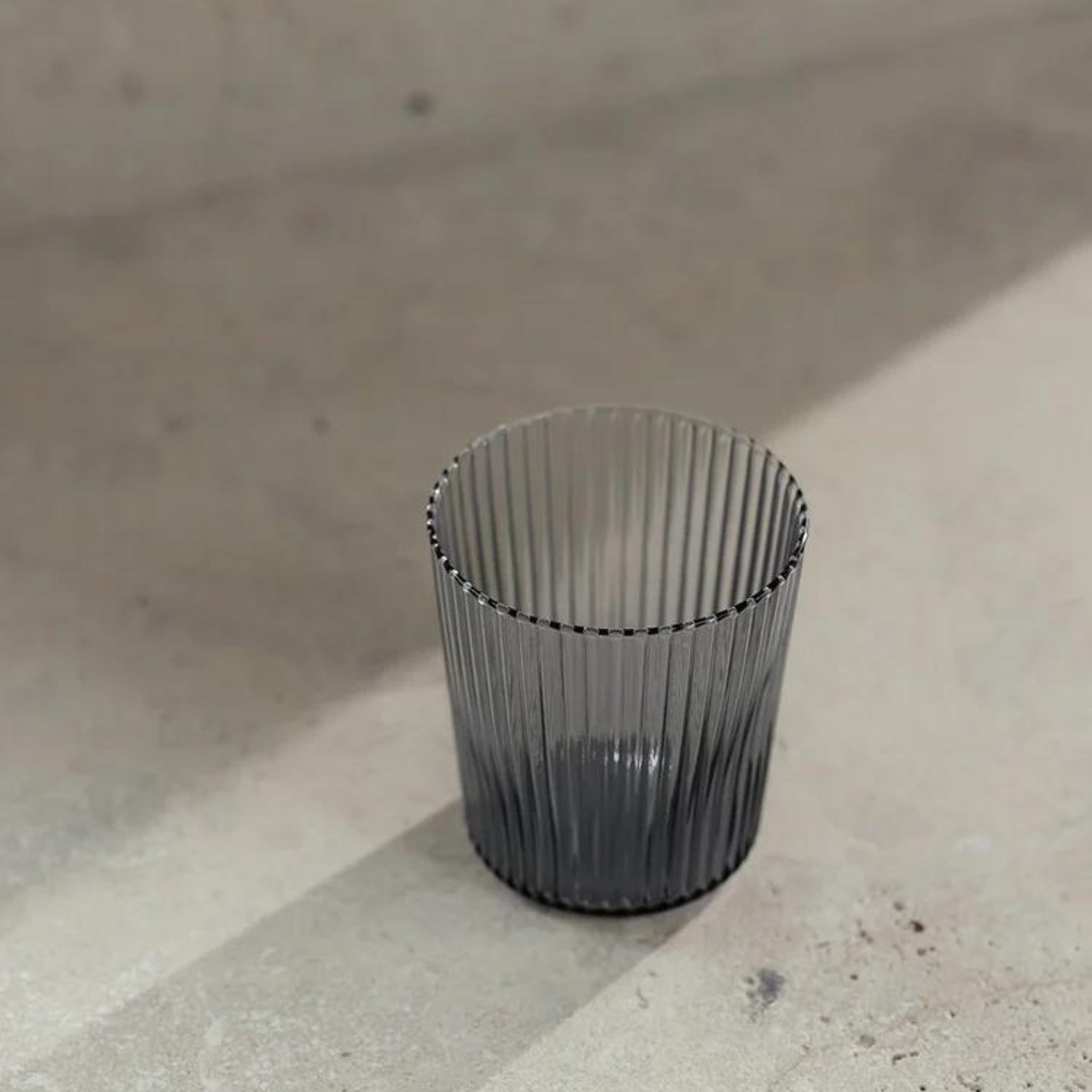 Een transparant, verticaal geribbeld Paveau Waterglas Twilight Blue van Paveau staat op een zonovergoten beige oppervlak en werpt een zachte schaduw - perfect voor wie houdt van stijlvol en duurzaam glaswerk.