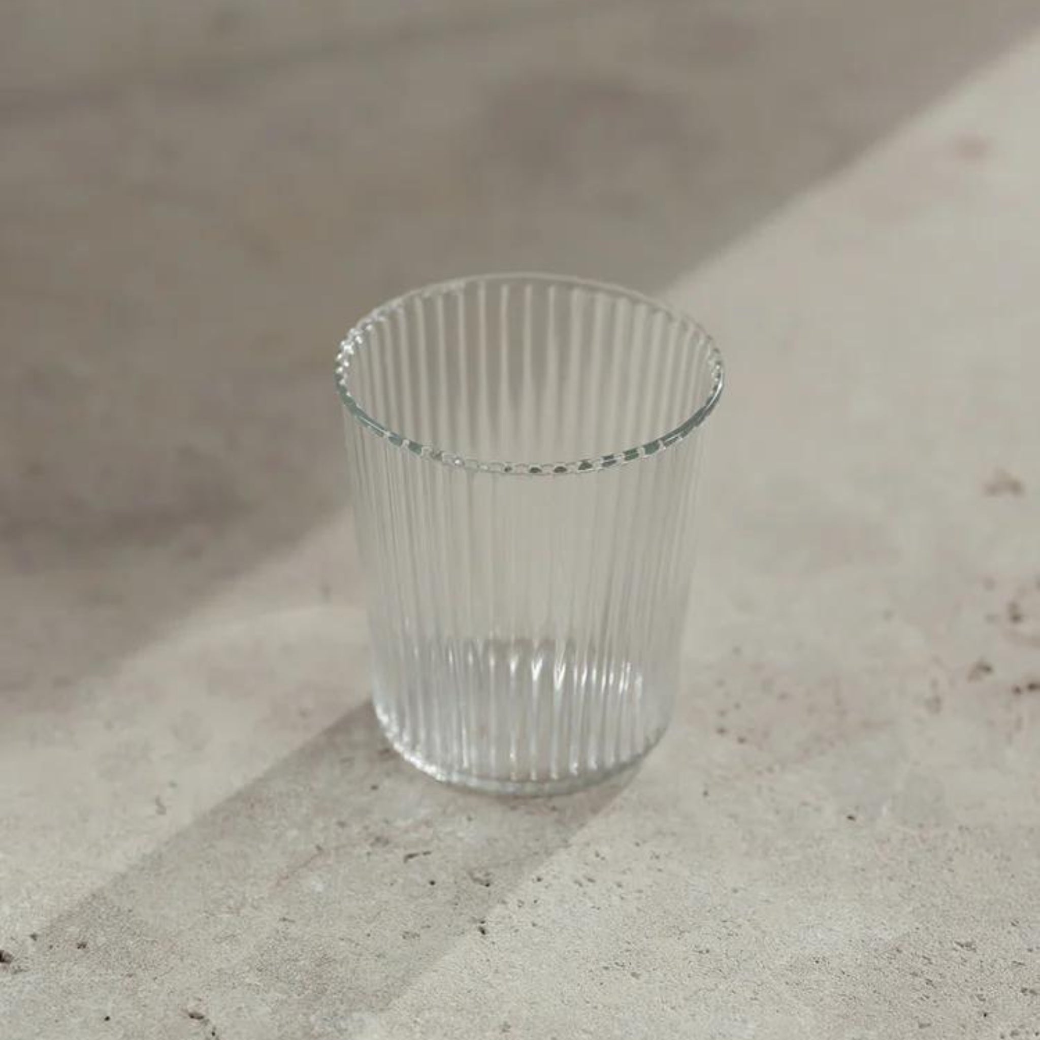 De Paveau Waterglas White Haven, met een geribbeld ontwerp, rust leeg op een licht oppervlak en werpt zachte schaduwen in natuurlijk zonlicht.