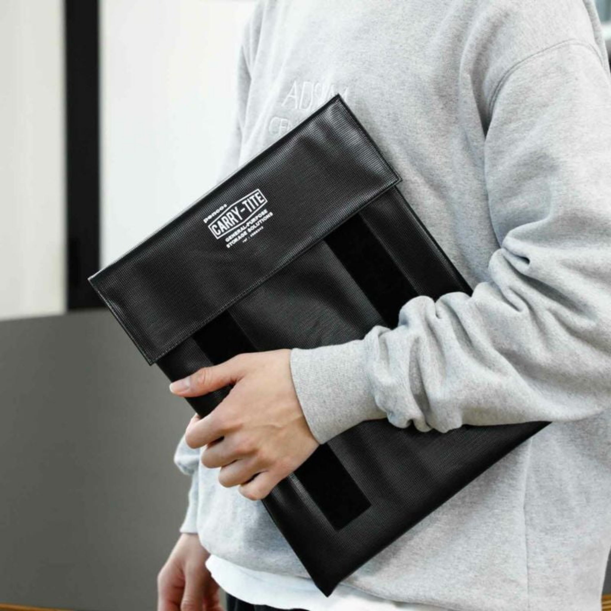 Een persoon in een grijs sweatshirt houdt een zwarte Penco Carry Tite Sleeve Zwart L tegen zijn zij. Deze hoes is gemaakt van waterdicht zeildoek en er passen laptops of documenten van 13 inch veilig en stijlvol in.