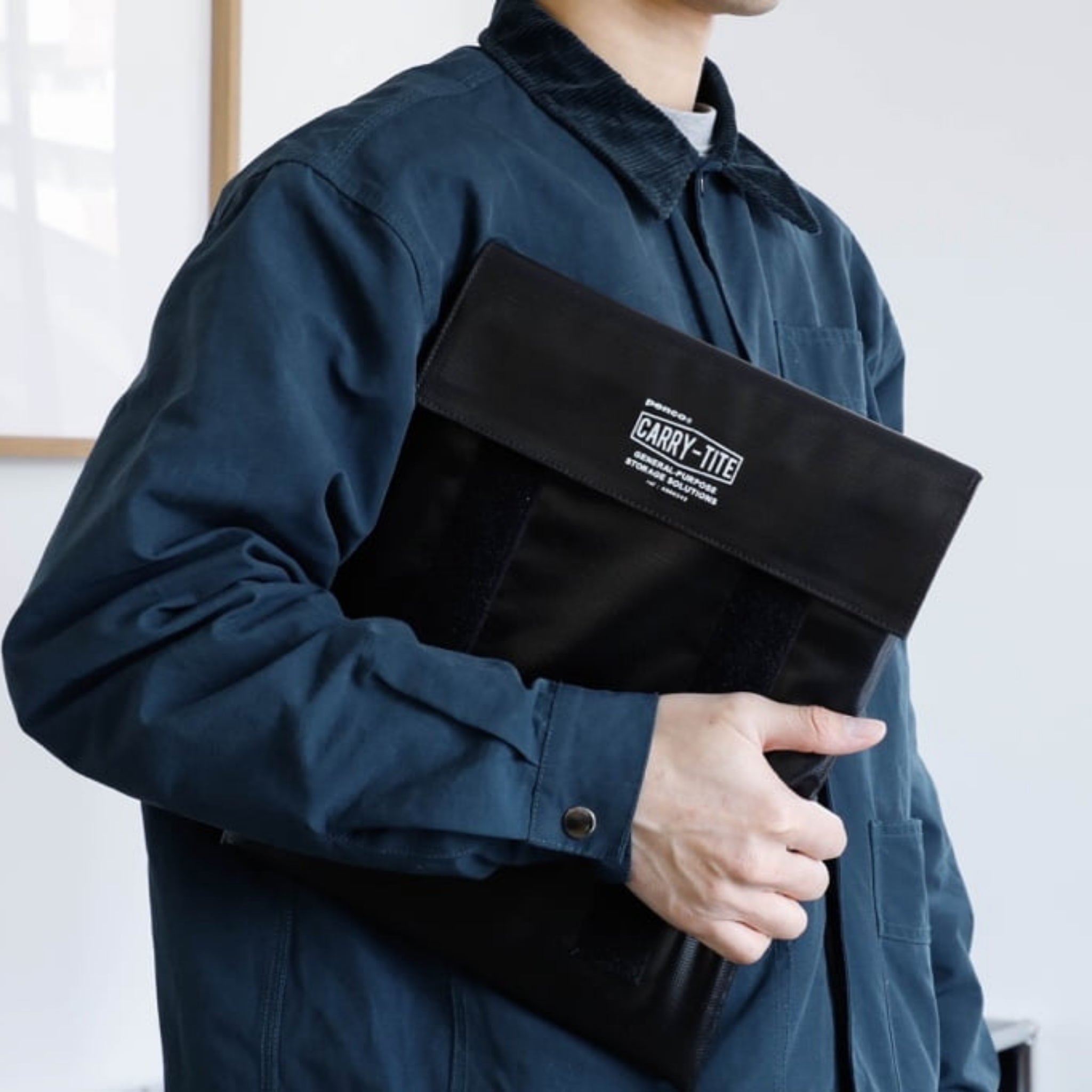 Een persoon in een blauwe jas houdt een Penco Carry Tite Sleeve Zwart L, gemaakt van waterdicht zeildoek en ideaal voor een 13-inch laptop, onder zijn arm terwijl hij binnen staat tegen een effen achtergrond.