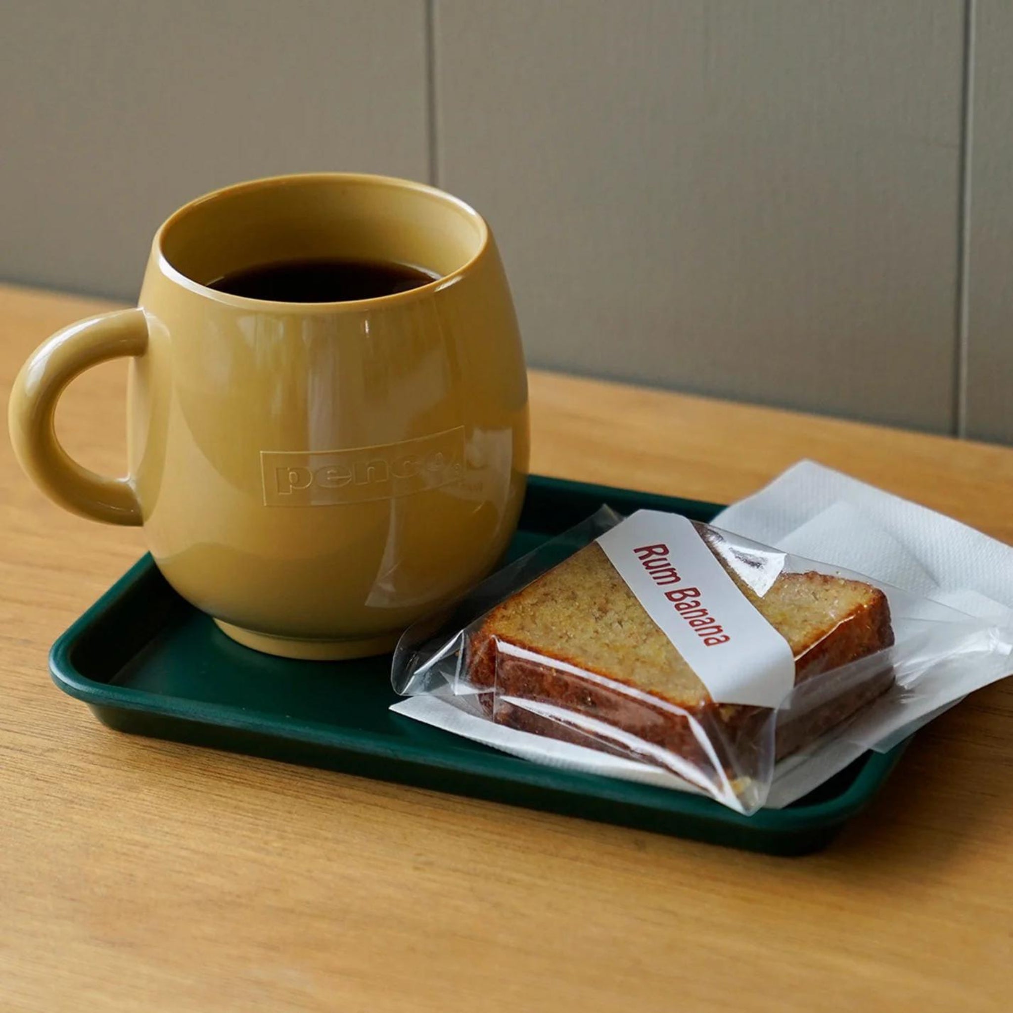 Een gele mok zwarte koffie staat op een dienblad van Penco Dienblad Donkergroen 18/12, naast verpakt bananenbrood met rum en een gevouwen wit servet, allemaal op een houten tafel.