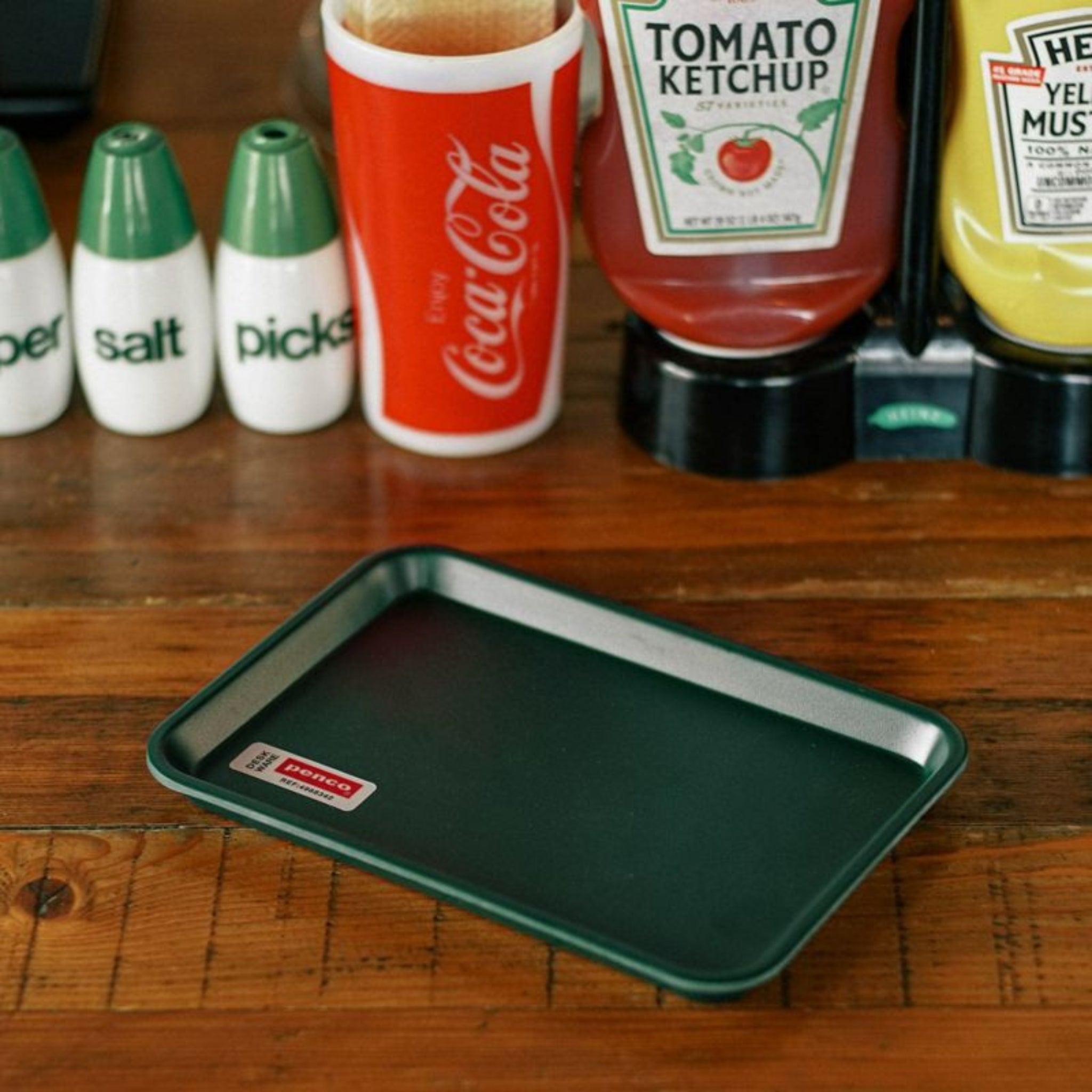 Een Dienblad Donkergroen 18/12 van Penco staat op een houten tafel, met op de achtergrond kruidenflesjes, een Coca-Cola beker en een groen peper- en zoutstel.