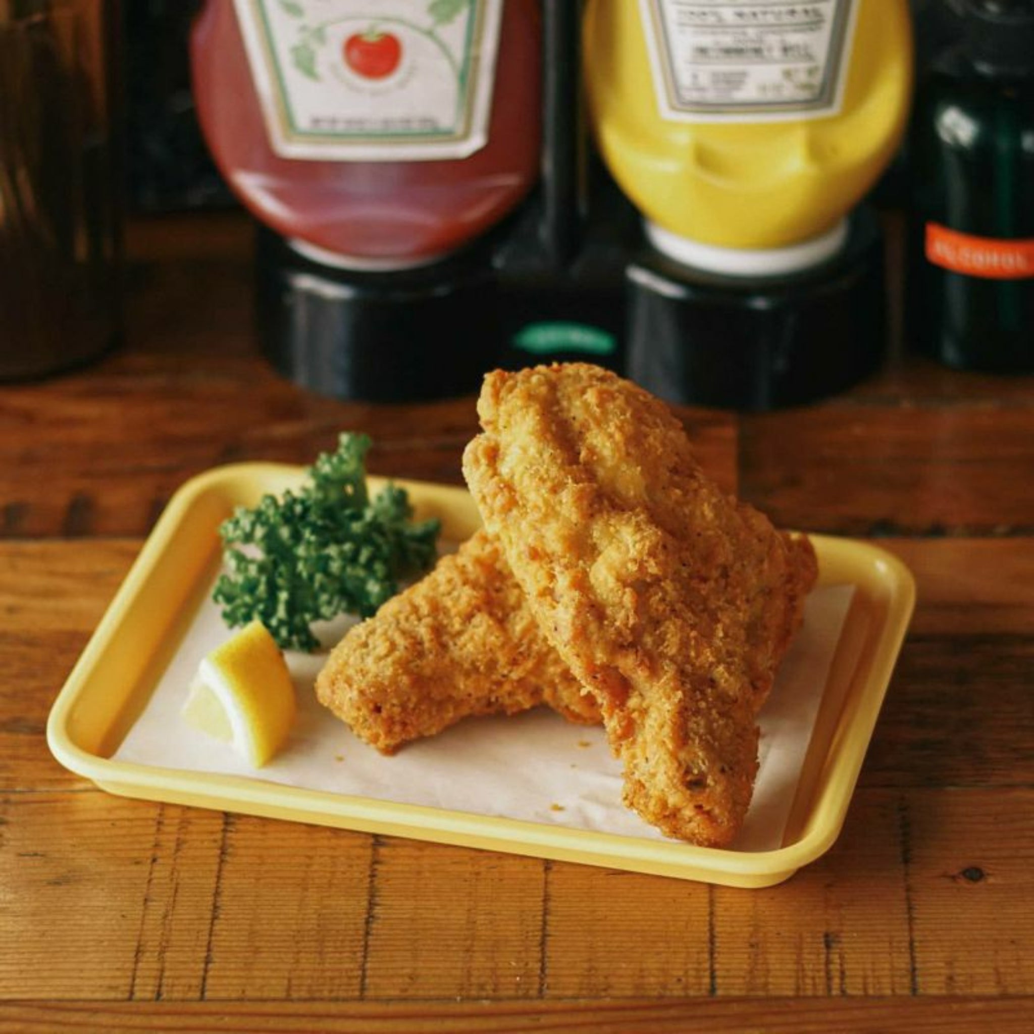 Een Penco Dienblad Geel 18/12 houdt twee stukken gebakken kip, een citroenpartje en peterselie vast. Op de achtergrond staan ketchup- en mosterdflessen op een houten oppervlak. Merk: Penco.