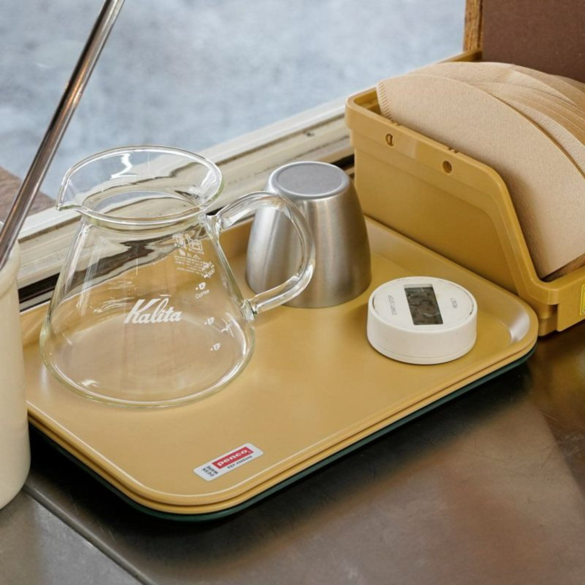 Een helder glazen Kalita koffieschep, een metalen maatbeker, een digitale timer en een bruine servettenhouder rusten op de Penco Dienblad Geel 28/21 (Penco) op een metalen aanrecht.