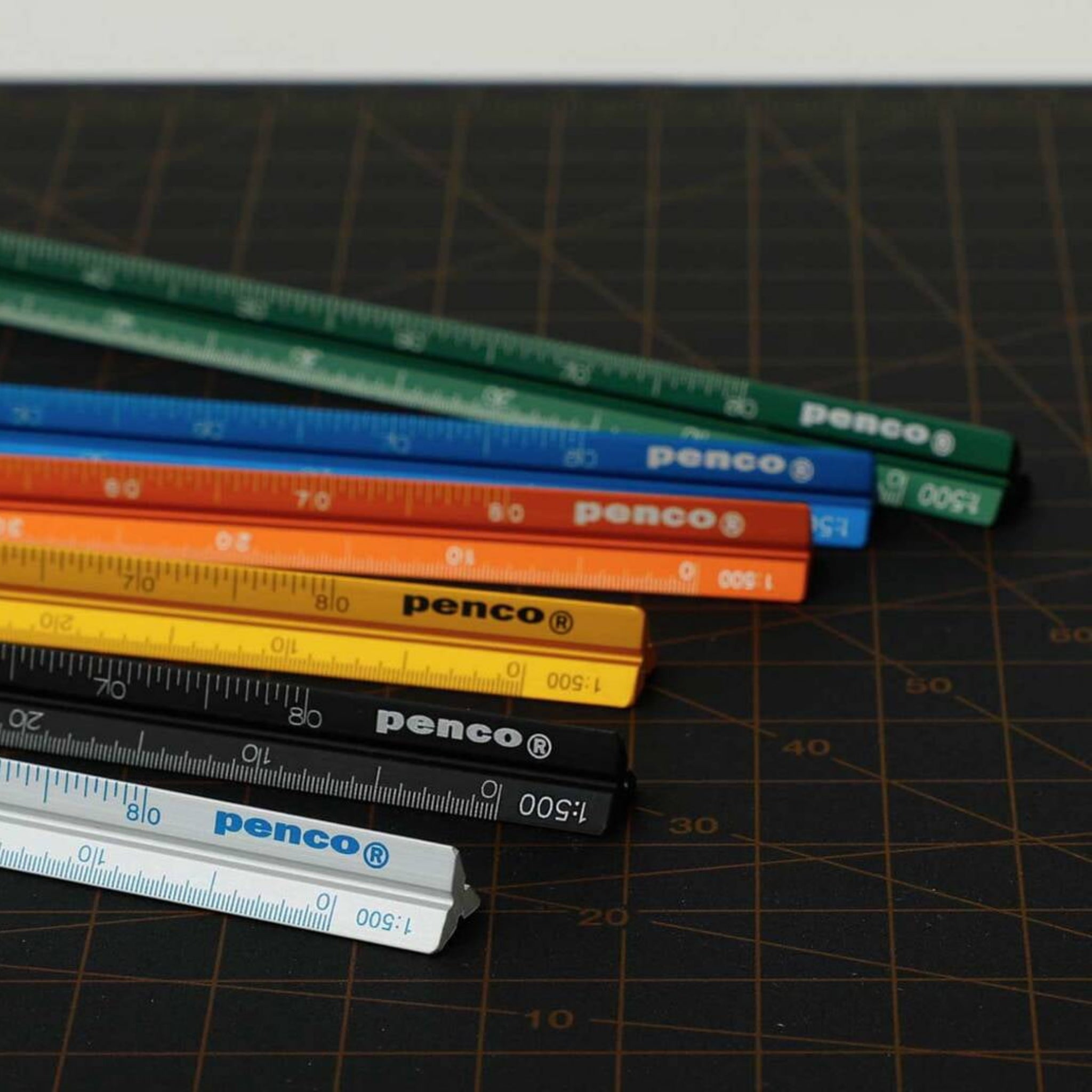 Zeven kleurrijke Penco Lat Aluminium Zilver driehoekige linialen, elk voorzien van een maatverdeling en het Penco logo, liggen in een waaiervorm op een donkere snijmat met rasterpatroon - perfect voor technisch tekenen.