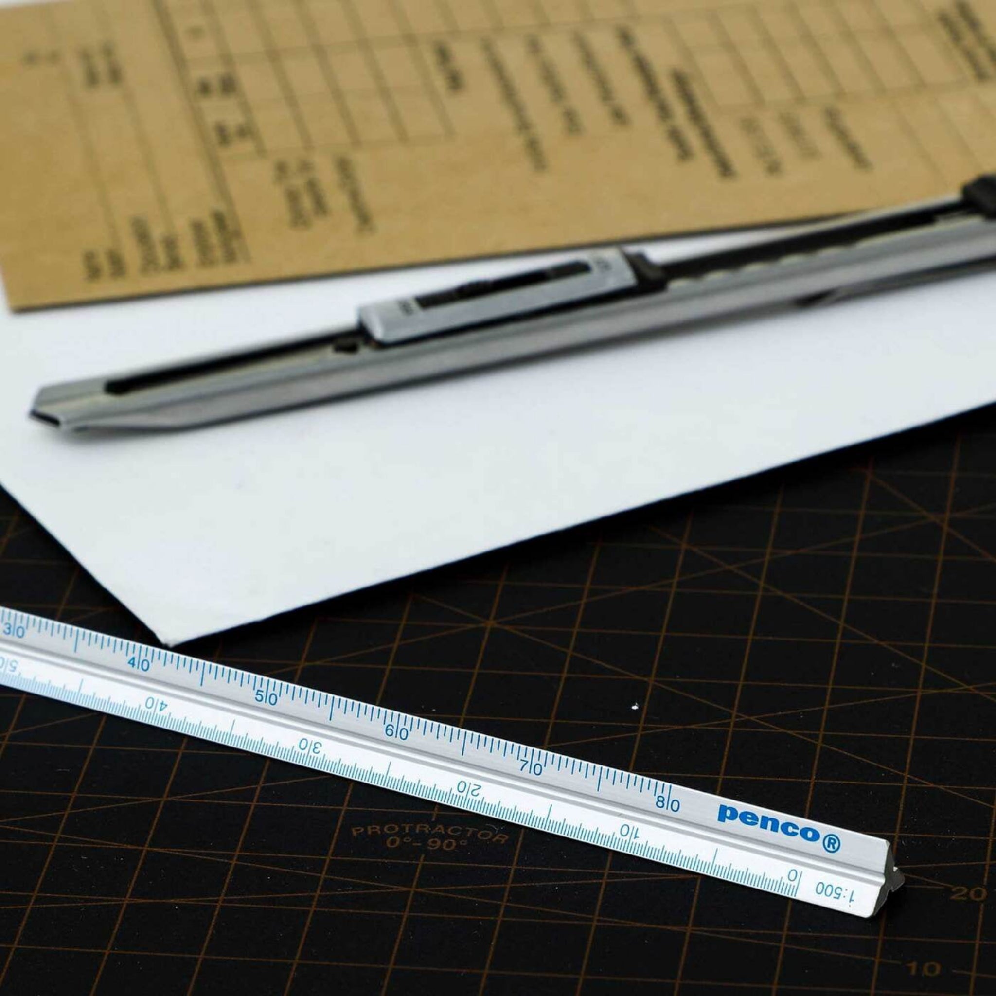 Een Penco Lat Aluminium Zilver, een hobbymes en vellen papier, waaronder een bruin formulier, liggen netjes op een donkere snijmat met maatlijnen - ideaal voor technische tekenklussen.