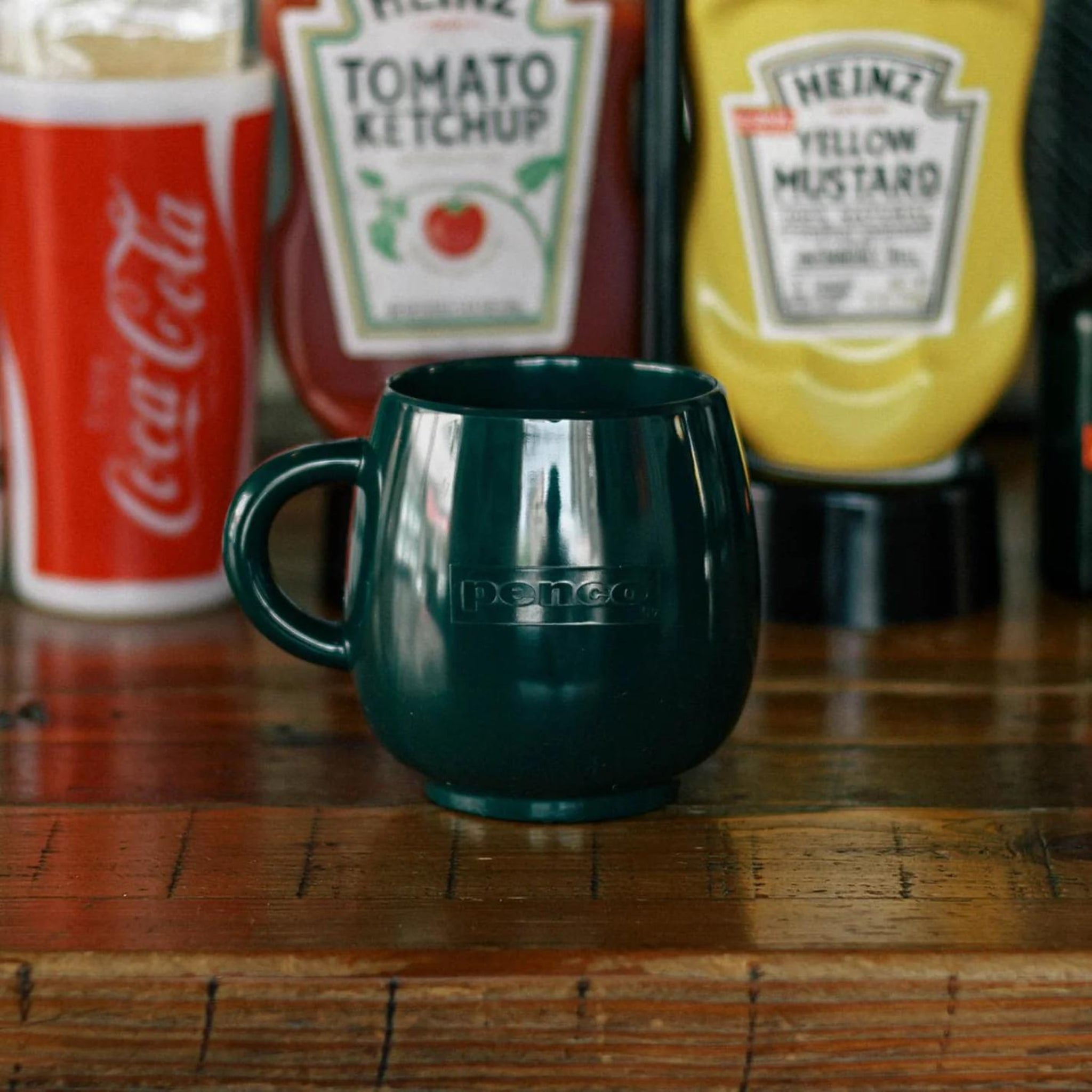 Een keramische mok van Penco Mok Donkergroen met een handvat en "Penco" in reliëf op de zijkant staat op een houten tafel voor ketchup- en mosterdflessen en een Coca-Cola beker.