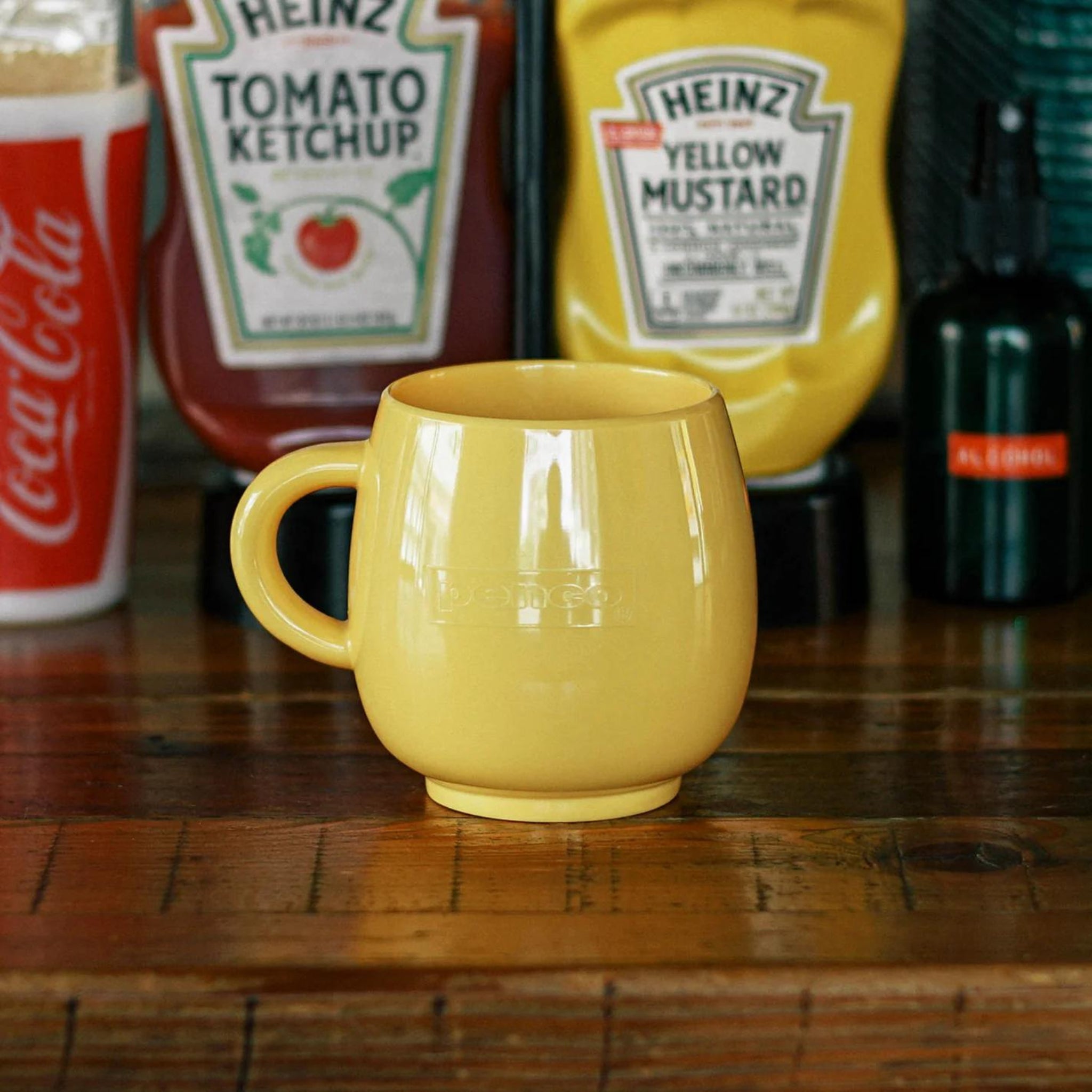 Een Penco Mok Geel staat op een houten tafel met Heinz tomatenketchup, Heinz gele mosterd, een rode Coca-Cola beker en een klein donker flesje op de achtergrond. Deze retro-mok van Penco voegt vintage charme toe aan het tafereel.