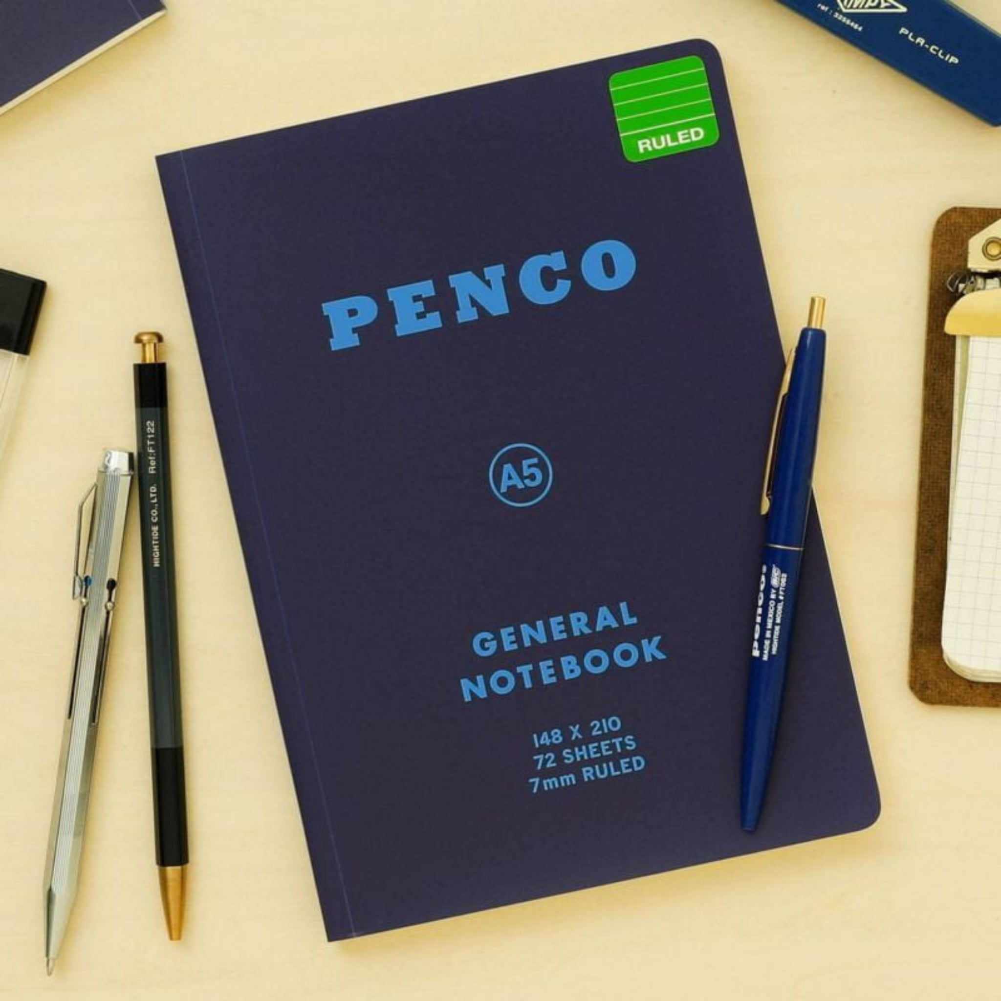 Een Penco Notitieboek Blauw A5 rust op een licht bureau met een pen, potlood, mechanisch potlood en clipboard-ideaal voor georganiseerd notitieboek werk. Blauwe tekst en een groene sticker geven dit donkerblauwe notitieboek extra stijl.