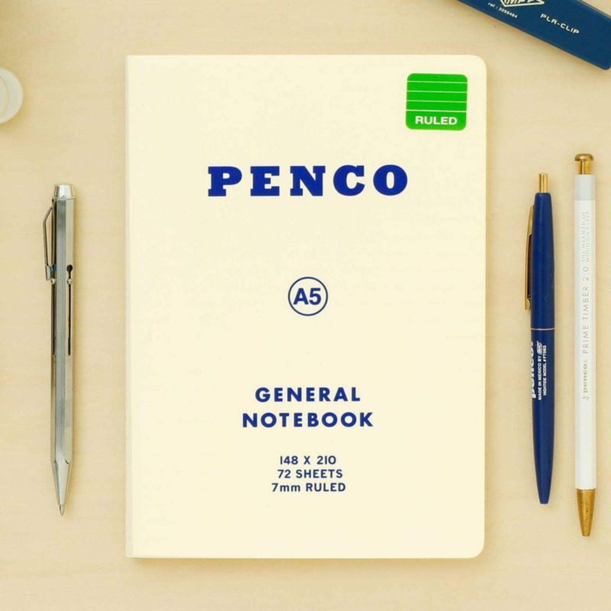 Een Penco Notitieboek Wit A5 met een groen gelijnd label ligt op een licht bureau, samen met een zilverkleurig vulpotlood, een blauwe pen en een wit-met-gouden pen - het perfecte notitieboek voor je werk.
