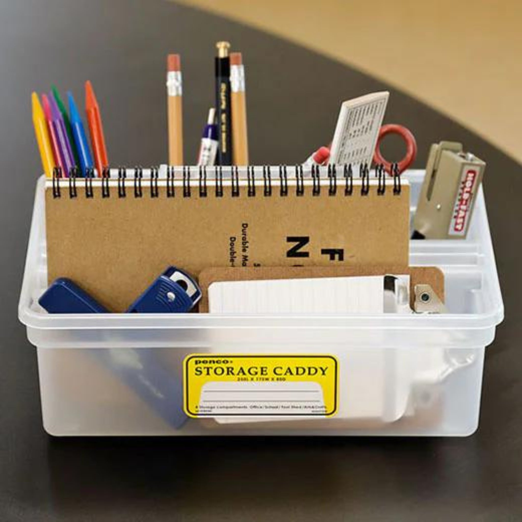 De Penco Opbergcaddy Helder van Penco organiseert gekleurde pennen, potloden, scharen, een nietmachine, notitieblok, indexkaarten en kantoorbenodigdheden. Het heeft een doorzichtig ontwerp met een geel "Storage Caddy" label op de voorkant.