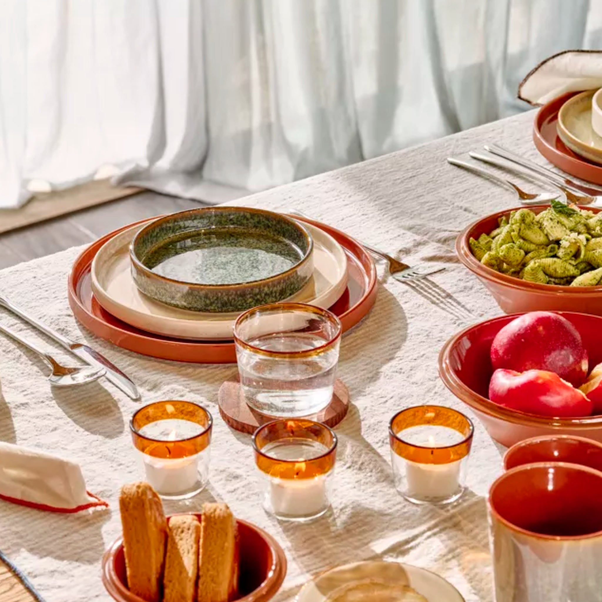 Op een zonovergoten eettafel staan borden Pomax Rasa Laag Bord Bruin D27, kommen met groene pasta en appels, amberkleurige waterglazen, broodstengels en kaarsen op een wit tafelkleed met textuur in de buurt van een raam met gordijnen.