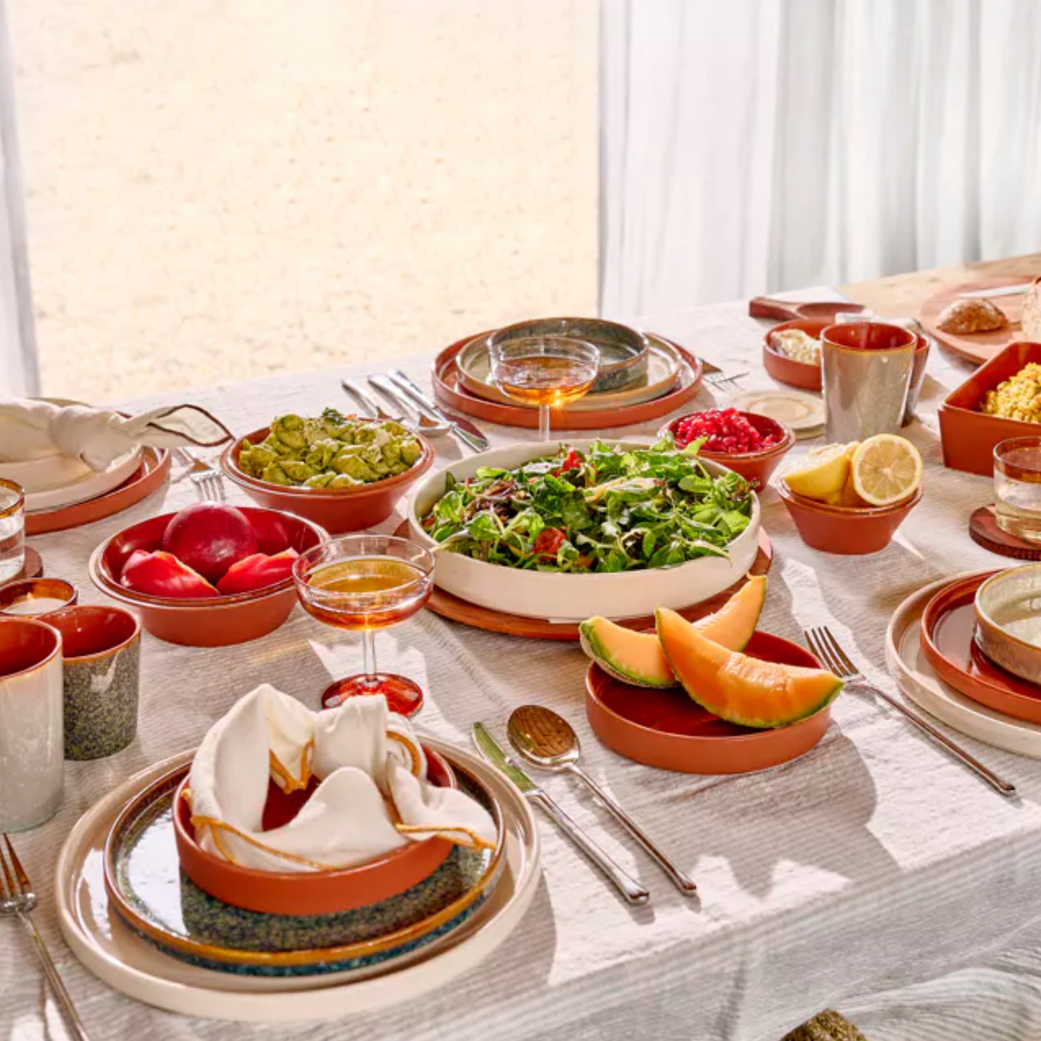 Een zonovergoten eettafel, gedekt met Pomax Rasa Kom Bruin D16 kommen, borden in aardetinten, salades, gesneden meloen, tomaten, brood, citroenen, sapglazen en schalen op een wit kleed - klaar voor de maaltijd.