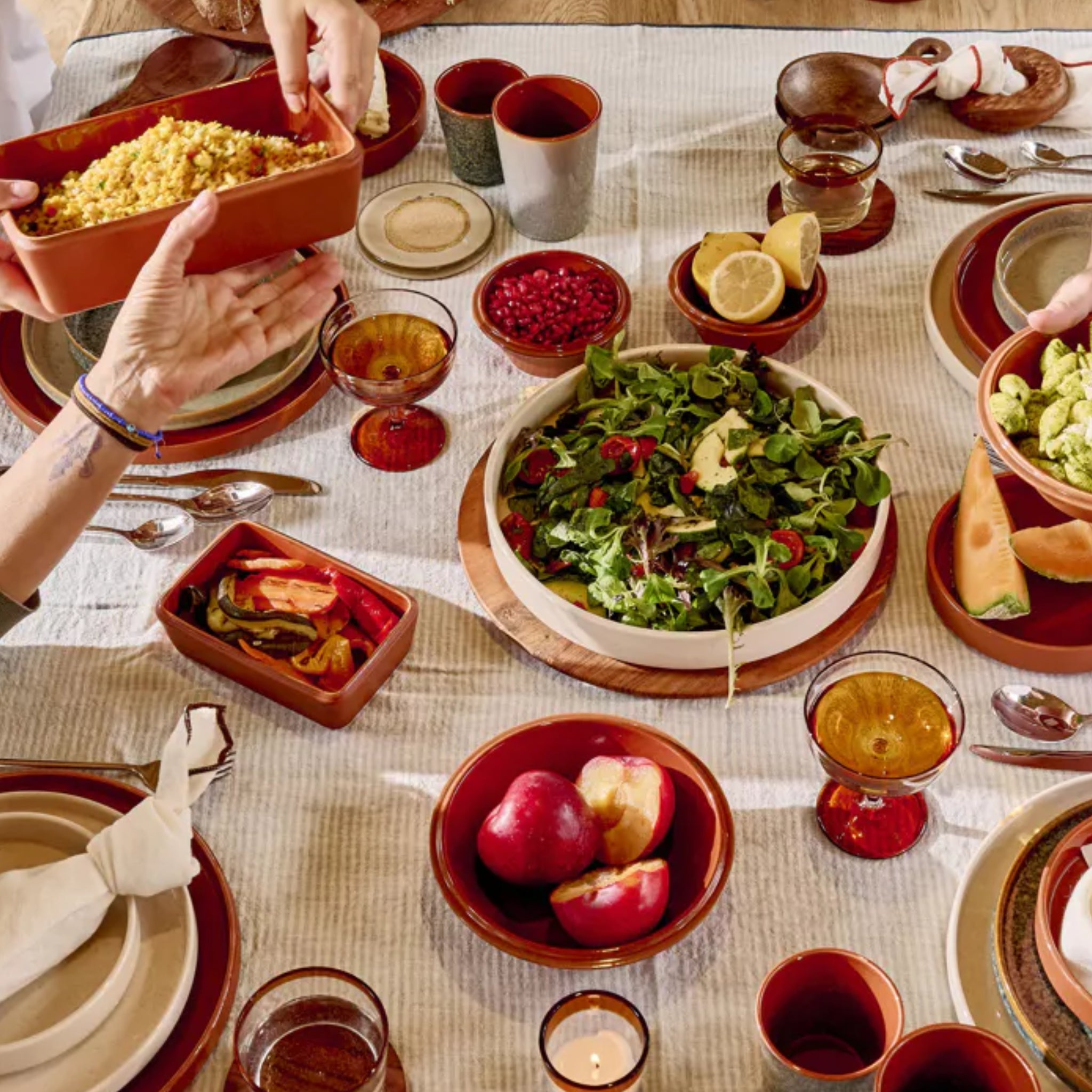 Een tafel is gedekt met verschillende gerechten op Pomax Rasa Laag Bord Bruin D27 borden, waaronder salade, fruit, granen en rode bessen, met drankjes in amberkleurige glazen. Handen serveren het eten, waardoor een warme sfeer ontstaat om de maaltijd te delen.
