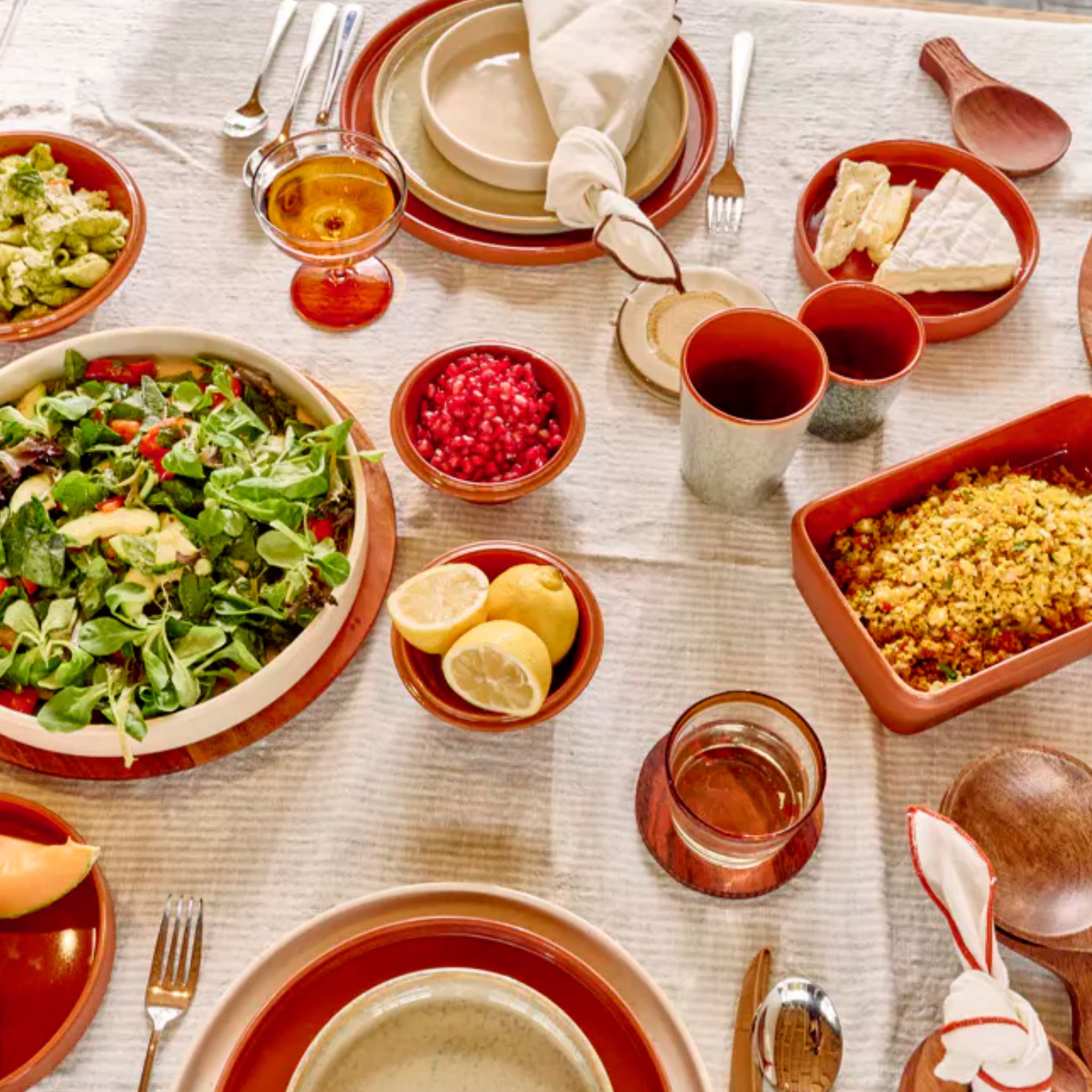 Een gedekte tafel met Pomax Rasa Laag Bord Bruin D27 borden toont salade, granaatappelpitjes, citroenhelften, avocado, brood en een ovenschotel als tafeldecoratie op een gestreept tafelkleed.