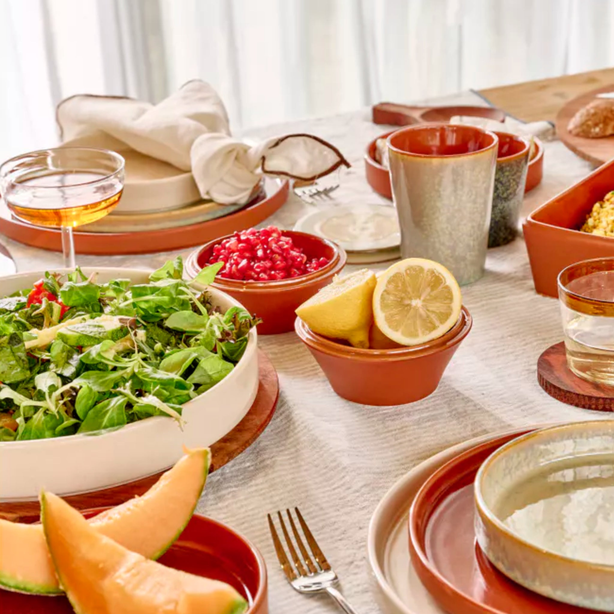 De Pomax Rasa Laag Bord Bruin D22 borden van Pomax zijn gedekt met salade, granaatappelpitjes, citroen, schijfjes meloen, brood en drankjes op een tafelkleed in aardetinten voor een informele tafeldecoratie en gastvrije eetsfeer.