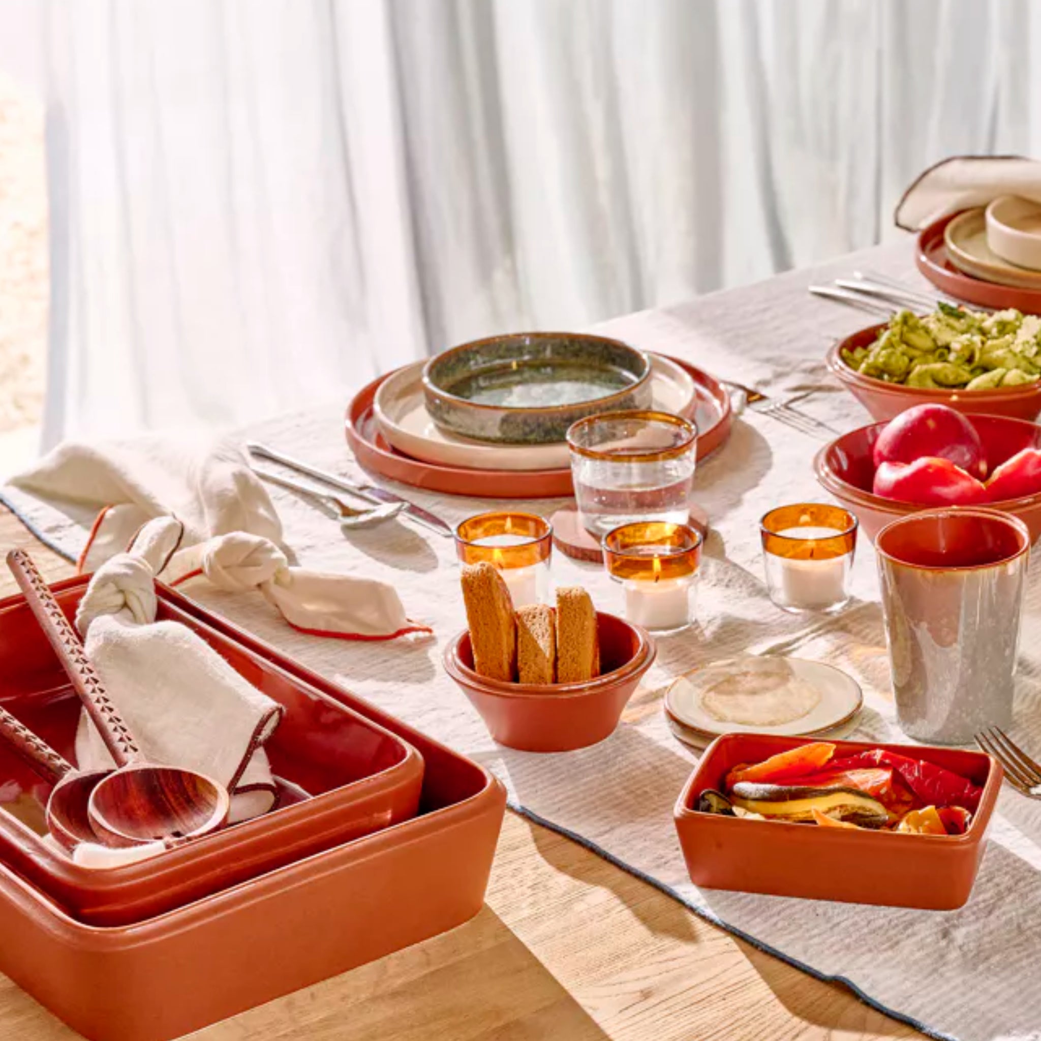 Een zonovergoten eettafel gedekt met Pomax Amuse Ovenschaal Bruin 39/24, porseleinen borden, groenteschalen, broodstengels, waterglazen en keukengerei op een licht kleed creëert een gezellige en uitnodigende maaltijdsetting.