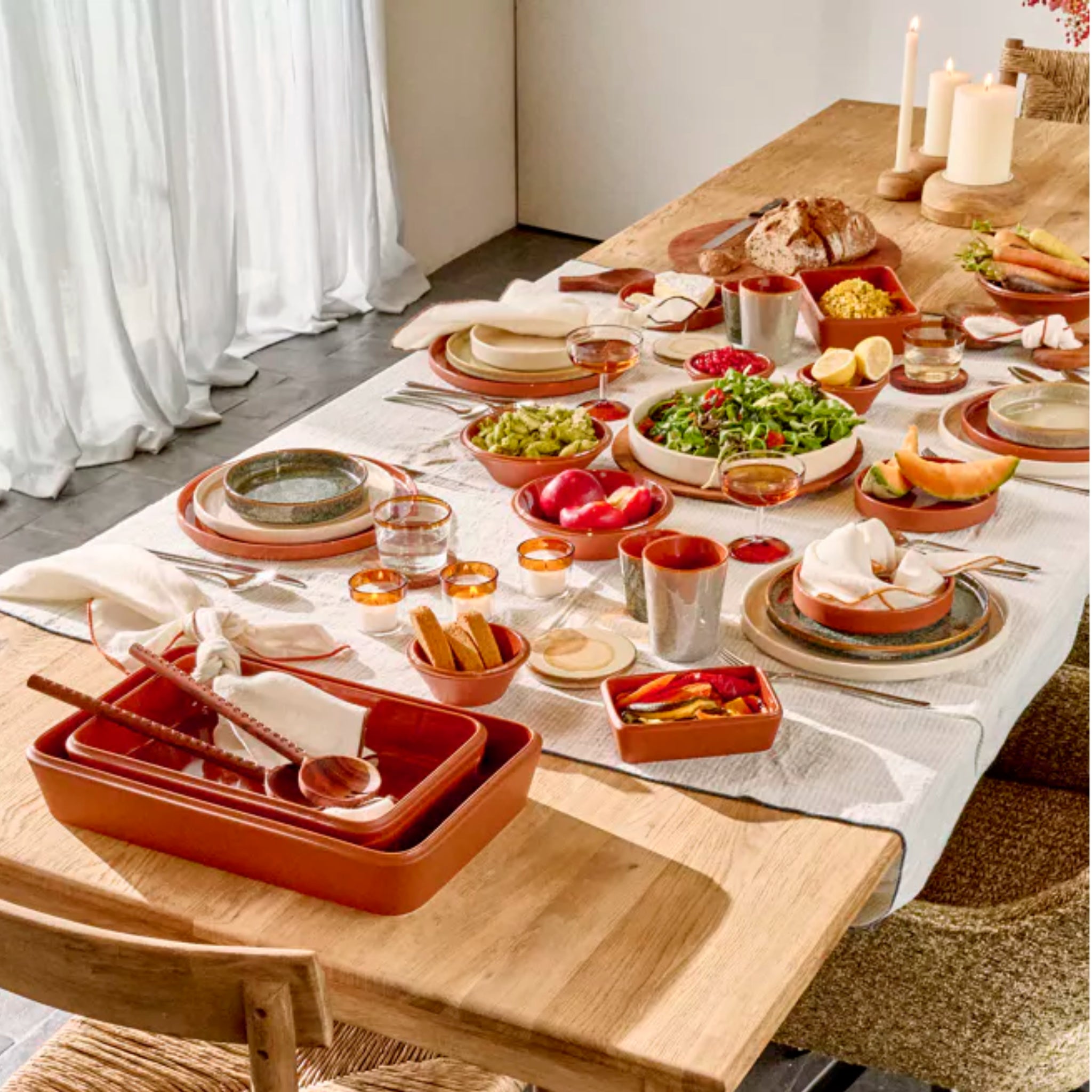 Een houten eettafel gedekt voor een maaltijd toont Pomax Amuse Ovenschaal Bruin 39/24, terracottakleurig servies, porseleinen borden, brood, kommen met groenten en zuiderse tafelaccessoires in natuurlijk licht door gordijnen.