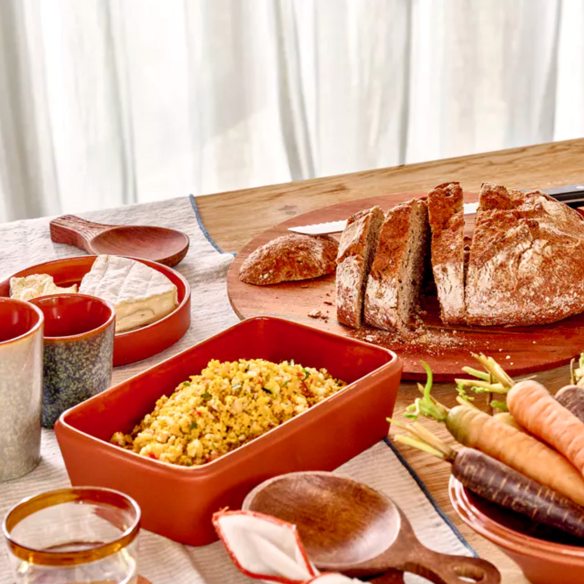 Een rustieke tafelschikking met gesneden rond brood op een Pomax Rasa Laag Bord Bruin D27, veelkleurige wortelen, couscous, brie kaas, keramische mokken en houten keukengerei op een licht tafelkleed met tafeldecoratie van Pomax.