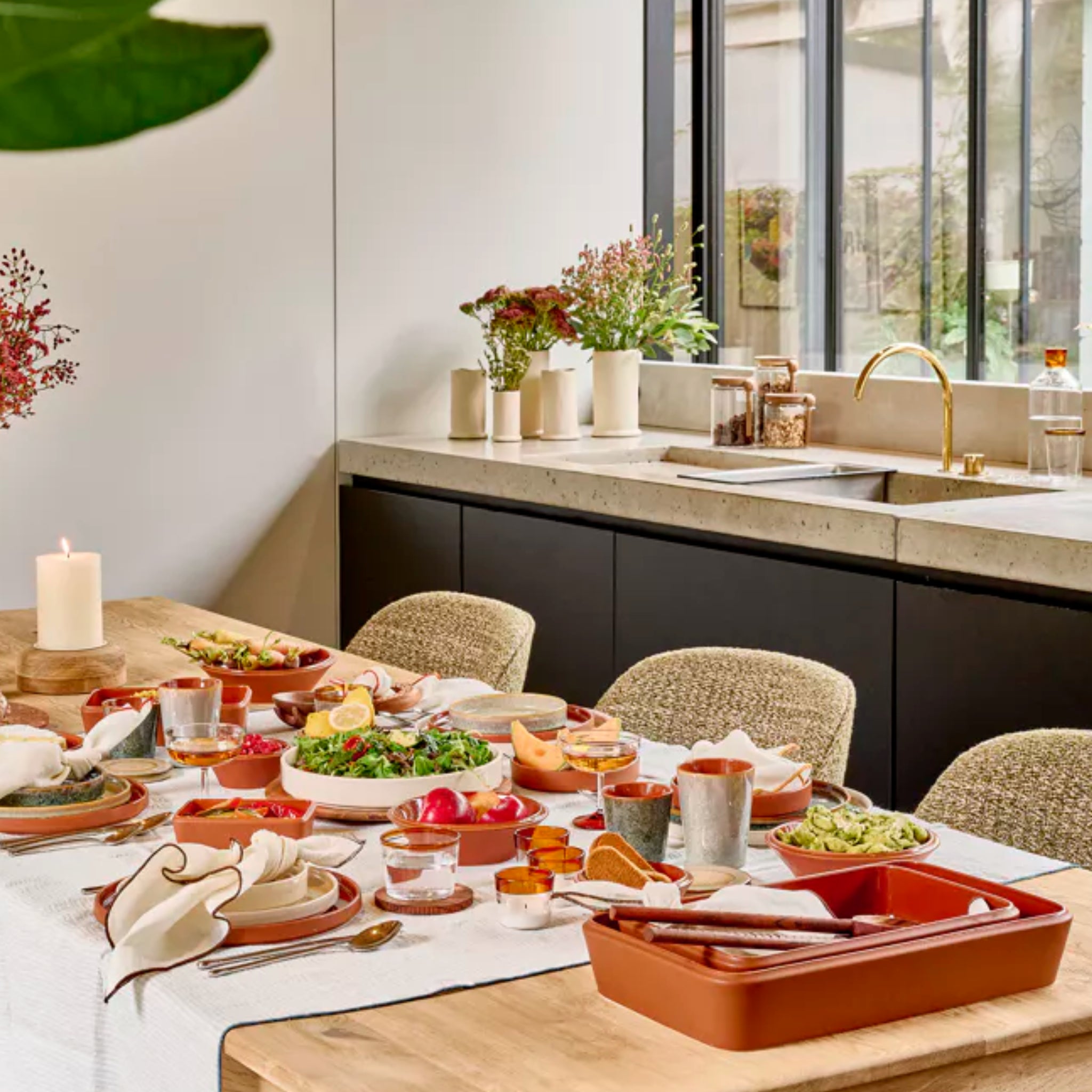 Een eettafel gedekt met porseleinen borden en de Pomax Amuse Ovenschaal Bruin 39/24, omringd door stoelen met kussens. Op de achtergrond is een moderne keuken te zien met een gootsteen, planten in potten en grote ramen.