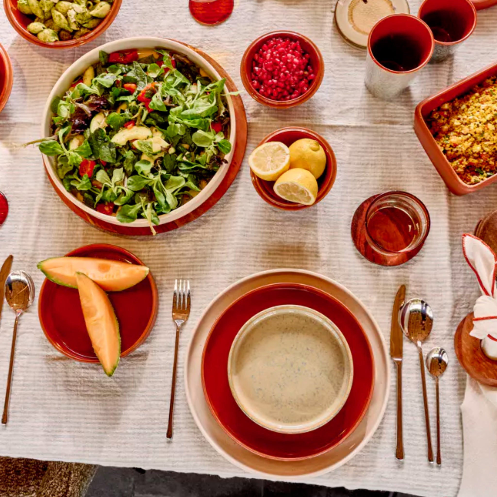 Een gedekte maaltijdtafel met Pomax Rasa Kom Wit D16 van Pomax in zachte aardetinten, met salade, citroenhelften, schijfjes cantaloupe, granaatappelpitjes en een drankje op een wit kleed met rood omrande borden en kommetjes.