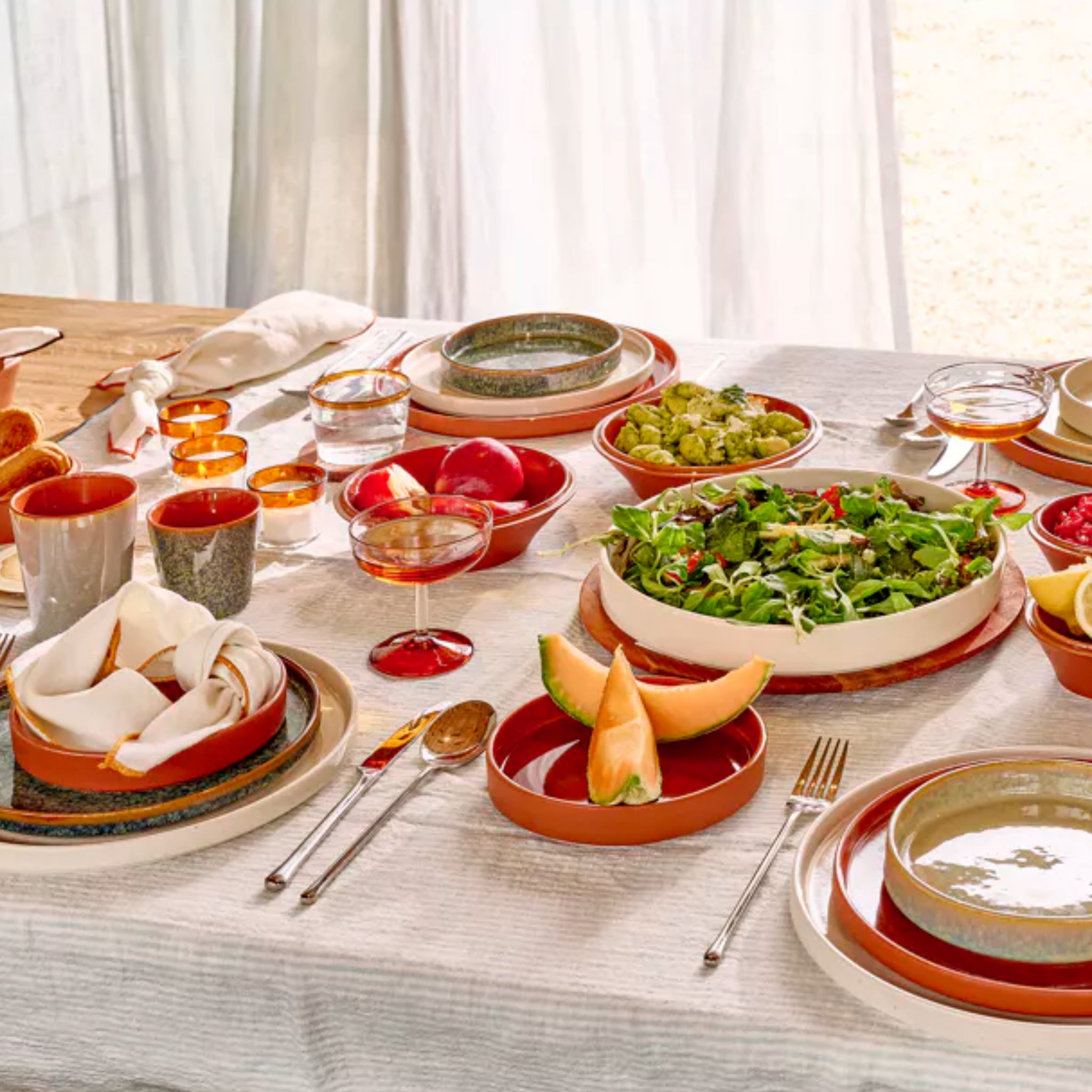 Een gedekte eettafel met Pomax Rasa Kom Bruin D16 van Pomax, aardetinten borden, salade, schijfjes meloen, avocado's, brood en drankjes. Het witte kleed en het natuurlijke licht creëren een warme en uitnodigende omgeving voor uw maaltijd.