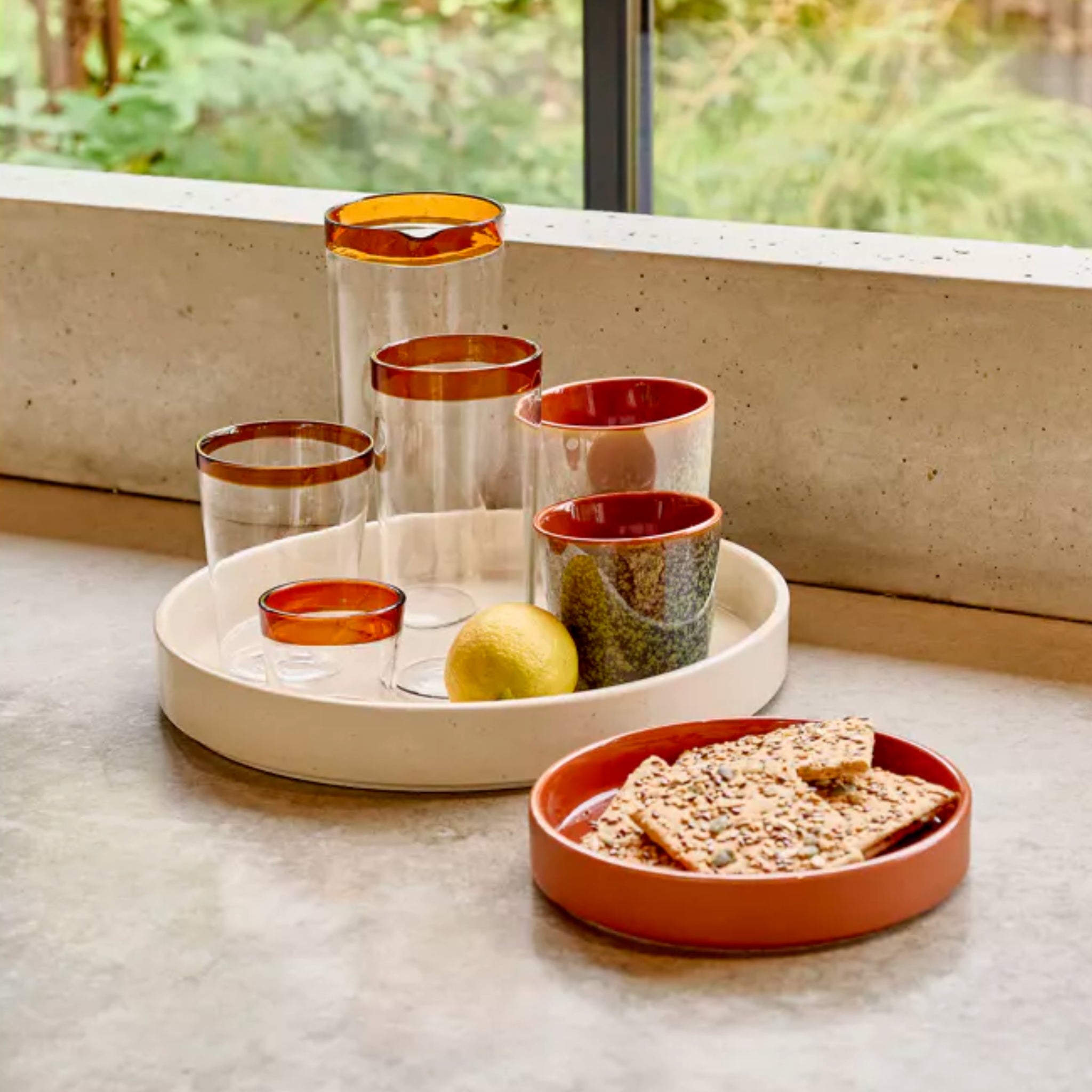 Op een rond dienblad staan vijf Pomax Rasa glazen met amberkleurige rand, een keramische mok en een citroen. Ernaast liggen crackers in de Pomax Rasa Kom Bruin D16-een warme tafeldecoratie met groen zichtbaar buiten.