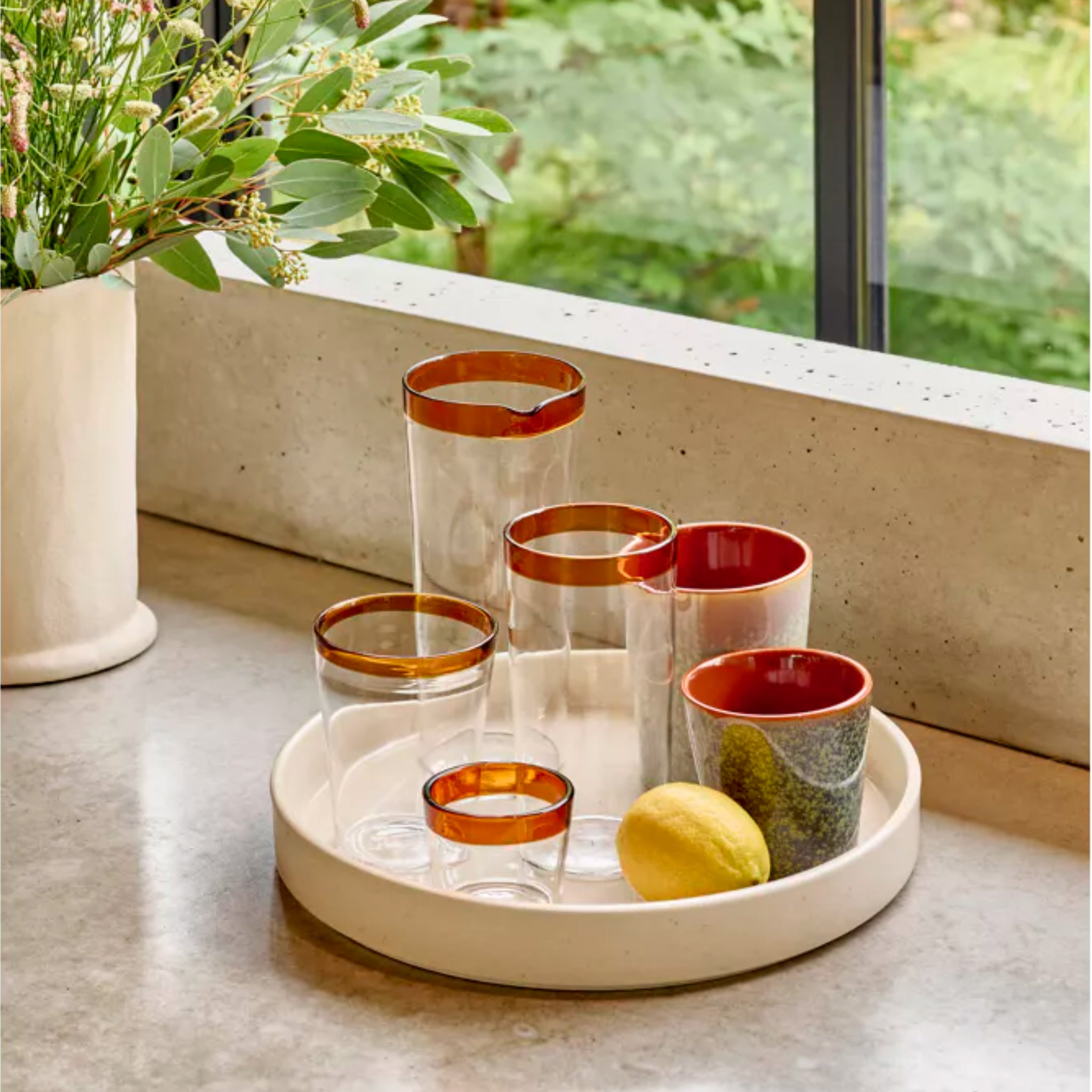 Een wit dienblad toont vijf heldere, vaatwasmachinebestendige glazen met bruine randen, twee Pomax Rasa Koffiekop Brons H8 kopjes en een citroen op een licht aanrecht in de buurt van groene bloemen-perfect tafeldecoratie van Pomax.