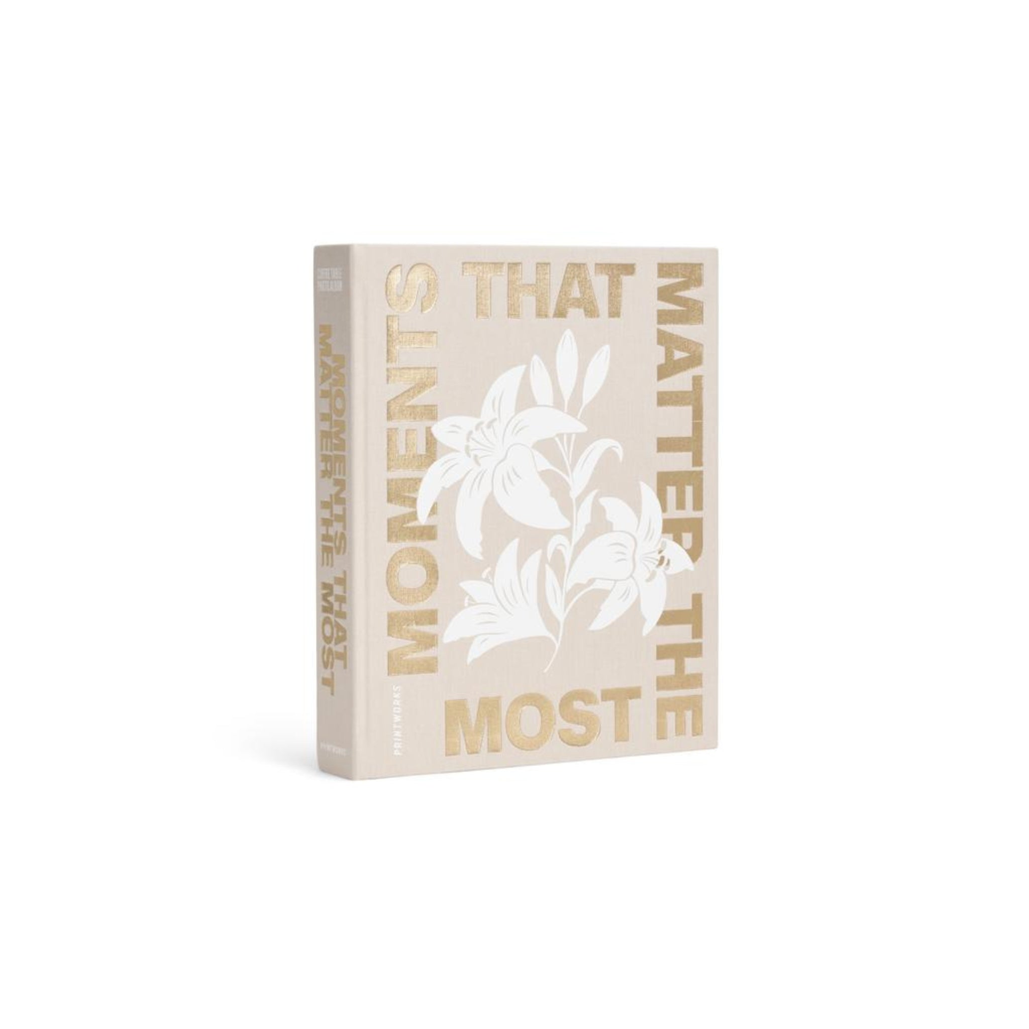 Het Printworks Fotoalbum Moments that Matter the Most is een beige hardcover koffietafelboek met gouden letters, een witte bloemillustratie op de omslag en de titel verticaal in goud op de rug - zowel stijlvol als tijdloos.