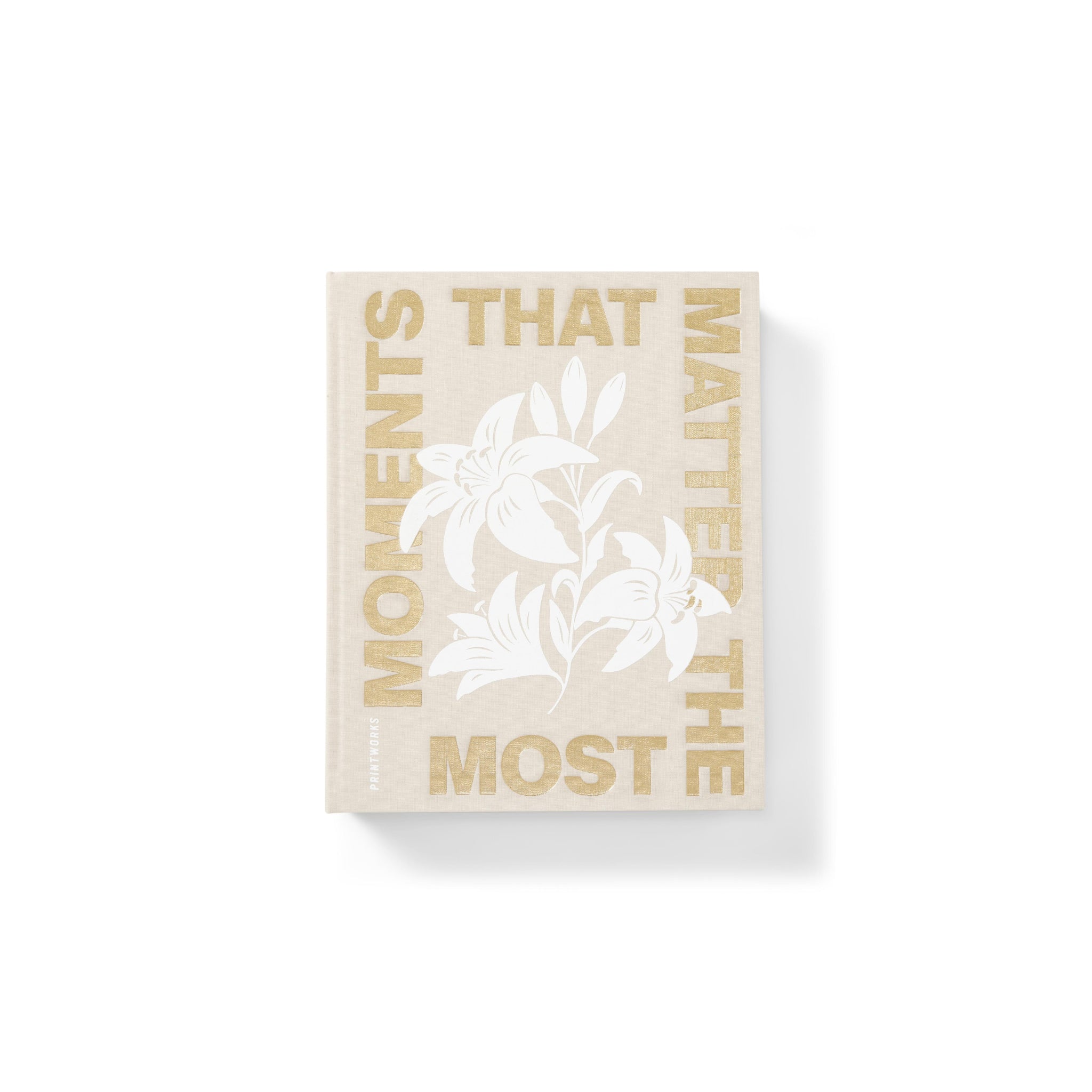 Het Printworks Fotoalbum Moments that Matter the Most heeft een beige hardcover met vetgedrukte gouden tekst en een witte bloemenillustratie, waardoor het een stijlvol salontafelaccent is. Gefotografeerd tegen een witte achtergrond.
