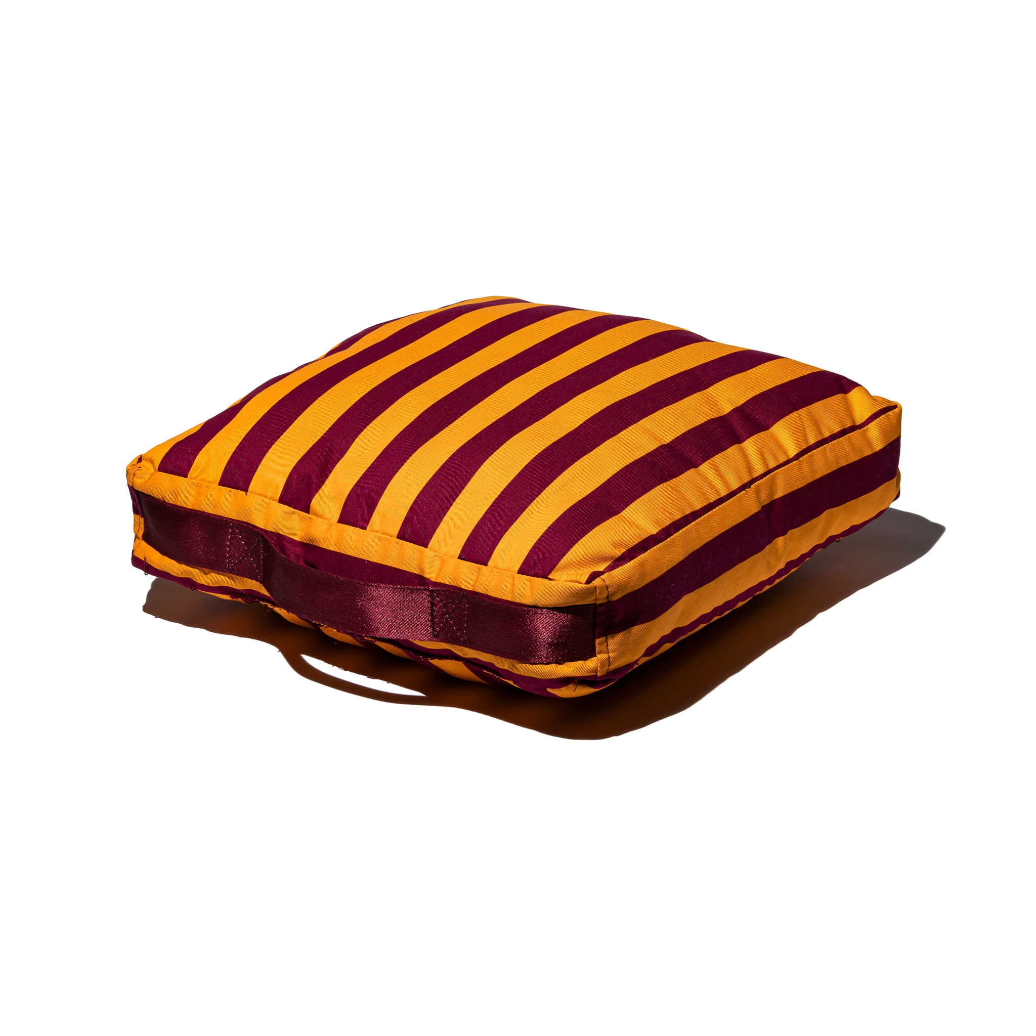 De Puebco Draagbaar Kussen Strepen Bordeaux x Mosterd is een vierkant kussen met bordeaux en mosterd strepen, plus een handig handvat aan de zijkant. Op een witte achtergrond met zachte schaduw voegt het extra zitcomfort toe aan elke ruimte.