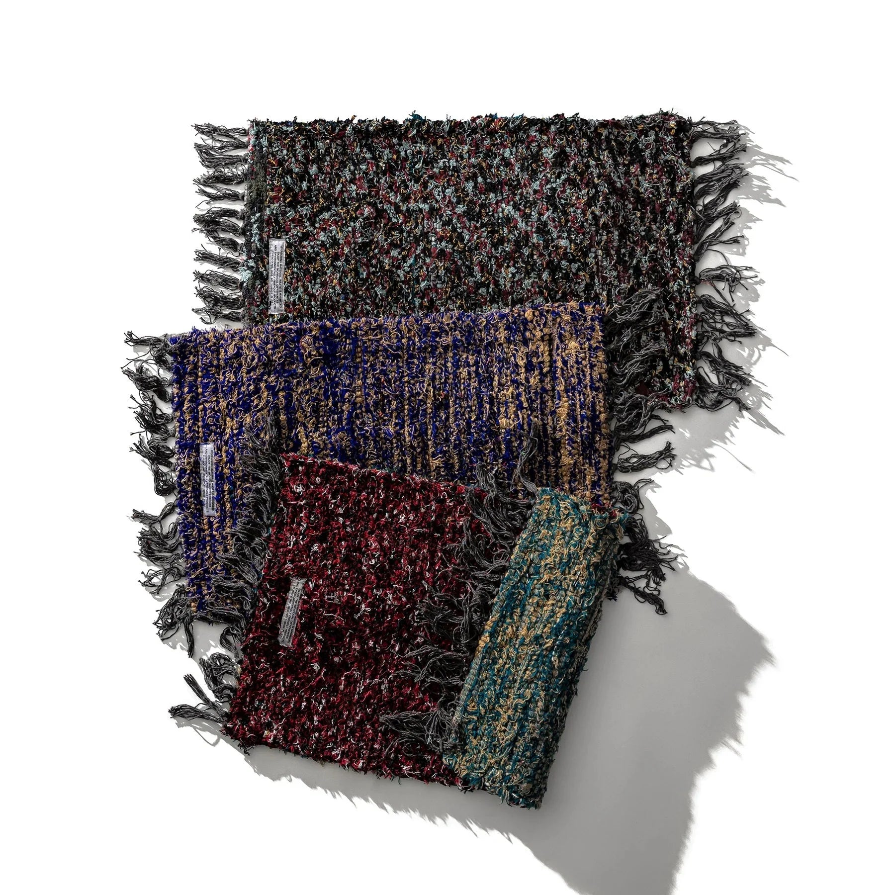 Drie veelkleurige geweven sjaals met textuur en franjes zijn diagonaal gerangschikt op een witte achtergrond, elk met een uniek patroon. Hun ambachtelijke stijl doet denken aan de Puebco Mat Gerecycleerd Textiel 63/41 van Puebco. Schaduwen naar rechts.