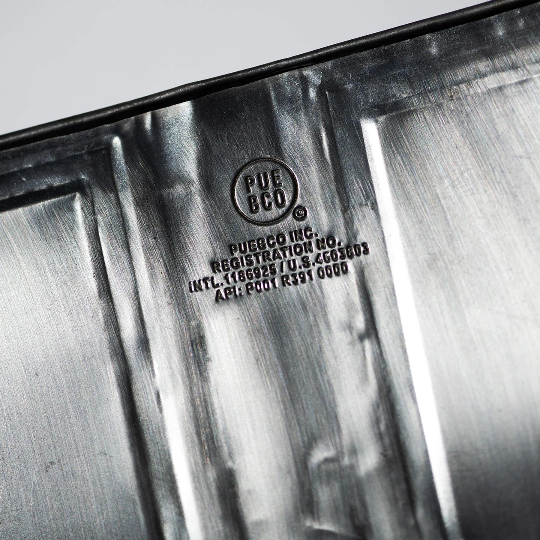 Close-up van een Puebco Prullenbak Gerecycleerd Staal H28: in reliëf aangebrachte lijnen en gestempelde tekst, waaronder PUEBCO INC., registratiegegevens en codes op het gerecyclede staal - ideaal voor industrieel gebruik. Merk: Puebco.