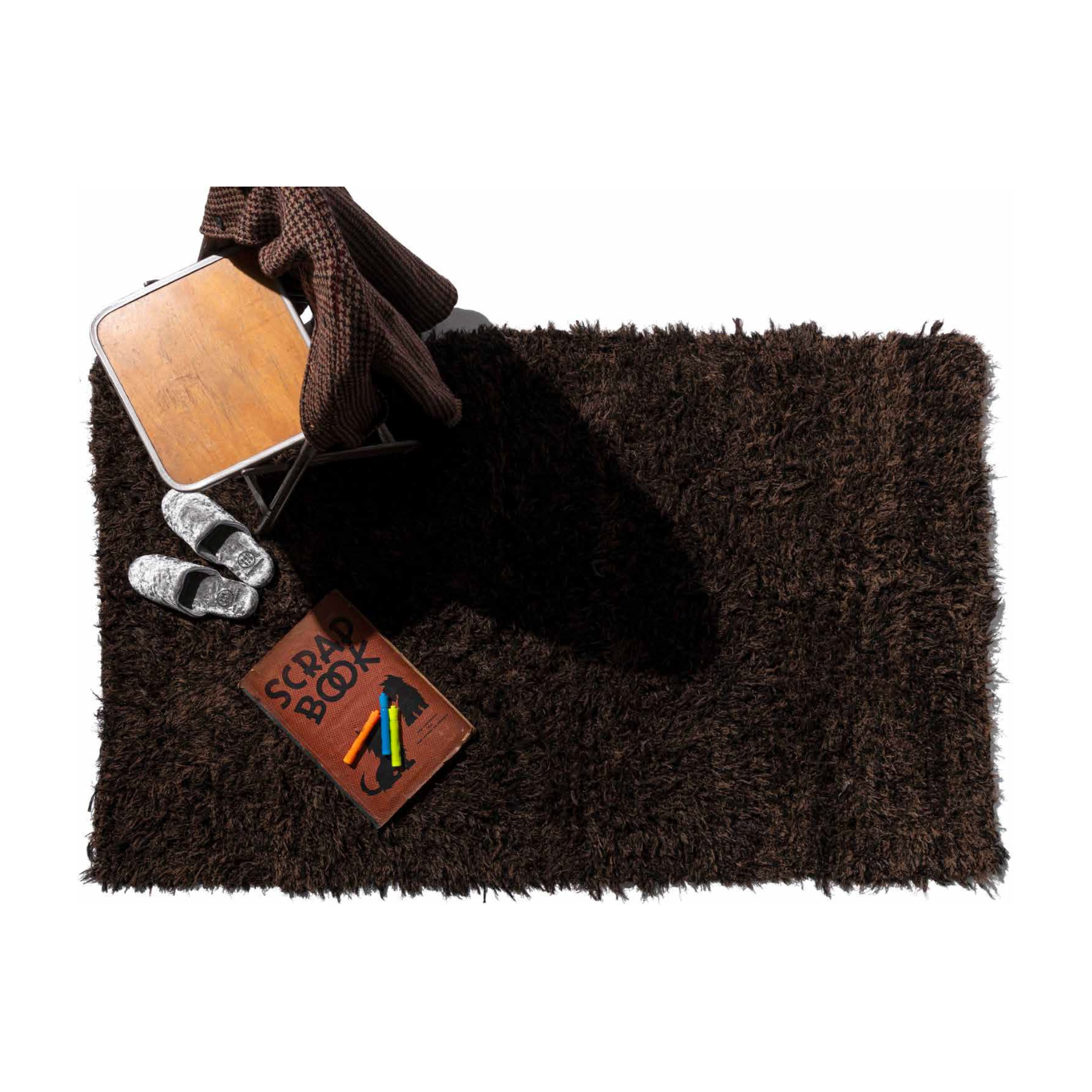 De Puebco Shaggy Tapijt Gerecycleerd Textiel 180/110 zit onder een houten stoel gedrapeerd in een bruin geruite deken, met grijze pantoffels in de buurt en een open plakboek met daarop twee gekleurde markers.