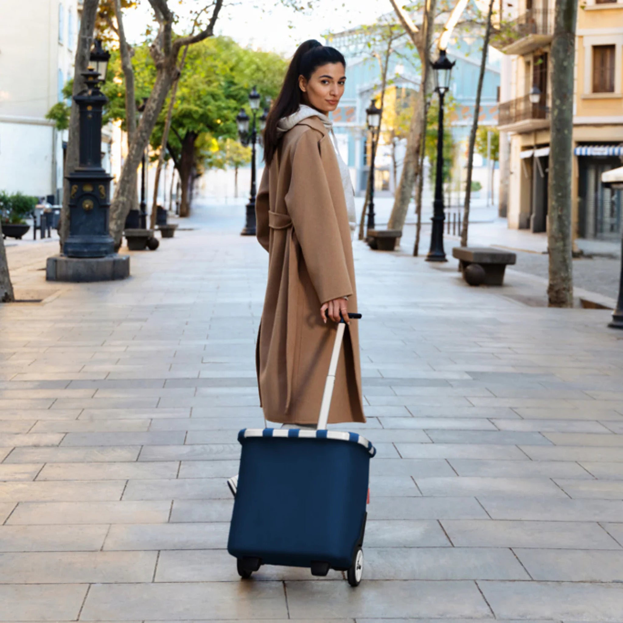 Een vrouw in een lange bruine jas loopt door een rustige, met bomen omzoomde stadsstraat, trekt een Reisenthel Carrycruiser Donkerblauw karretje en kijkt achterom over haar schouder.