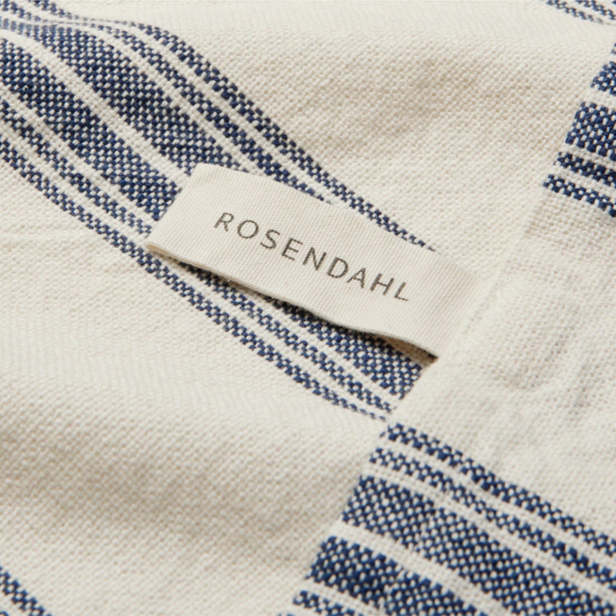 Een close-up van de Rosendahl Garn Theedoek Blauw 50/70 toont crèmekleurige stof met blauwe strepen en een wit Rosendahl-label-een elegant detail dat tijdloos Scandinavisch design belichaamt.