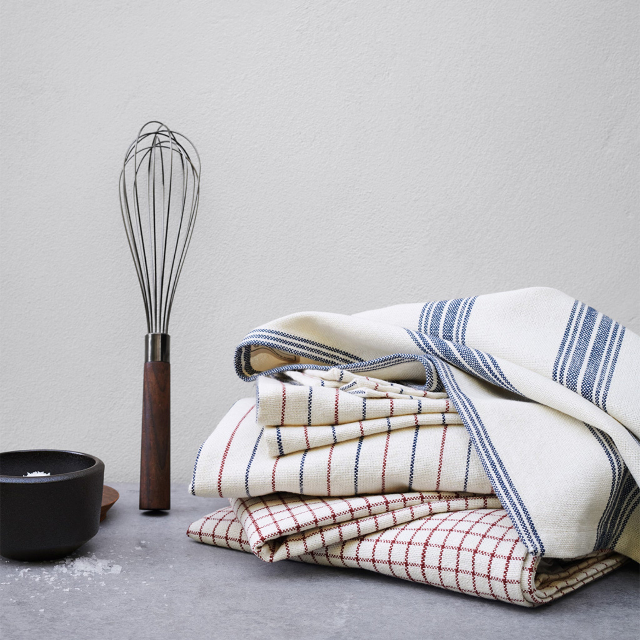 Een stapel Rosendahl Garn Theedoek Blauw 50/70 keukenhanddoeken met blauwe strepen ligt op een grijs oppervlak naast een metalen garde en een zwart schaaltje zout en geeft je keuken Scandinavische stijl. De achtergrond is een effen lichte muur.