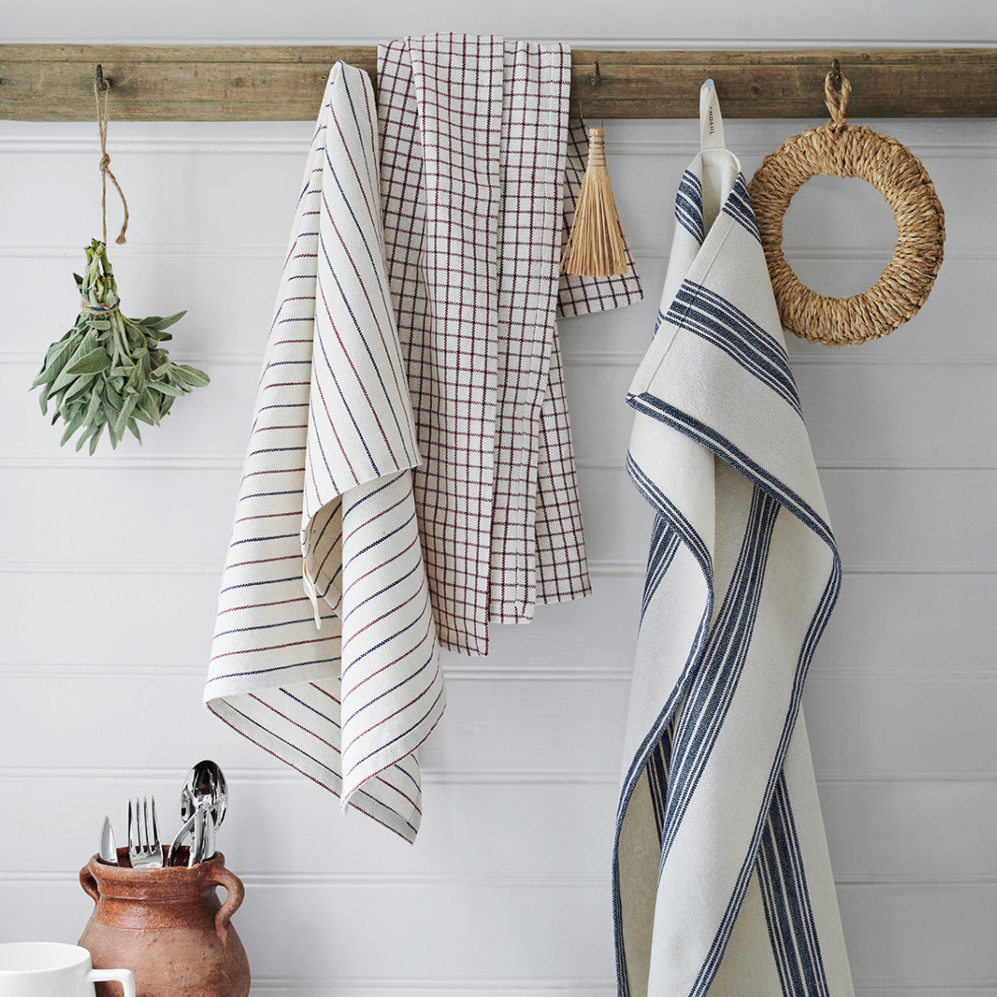Drie keukendoeken, waaronder de Rosendahl Garn Theedoek Blauw 50/70 van Rosendahl met gestreepte en geruite patronen, hangen aan een houten wasknijper naast kruiden, een kwastje, een geweven ring en een pot van klei op een wit gelambriseerde muur.