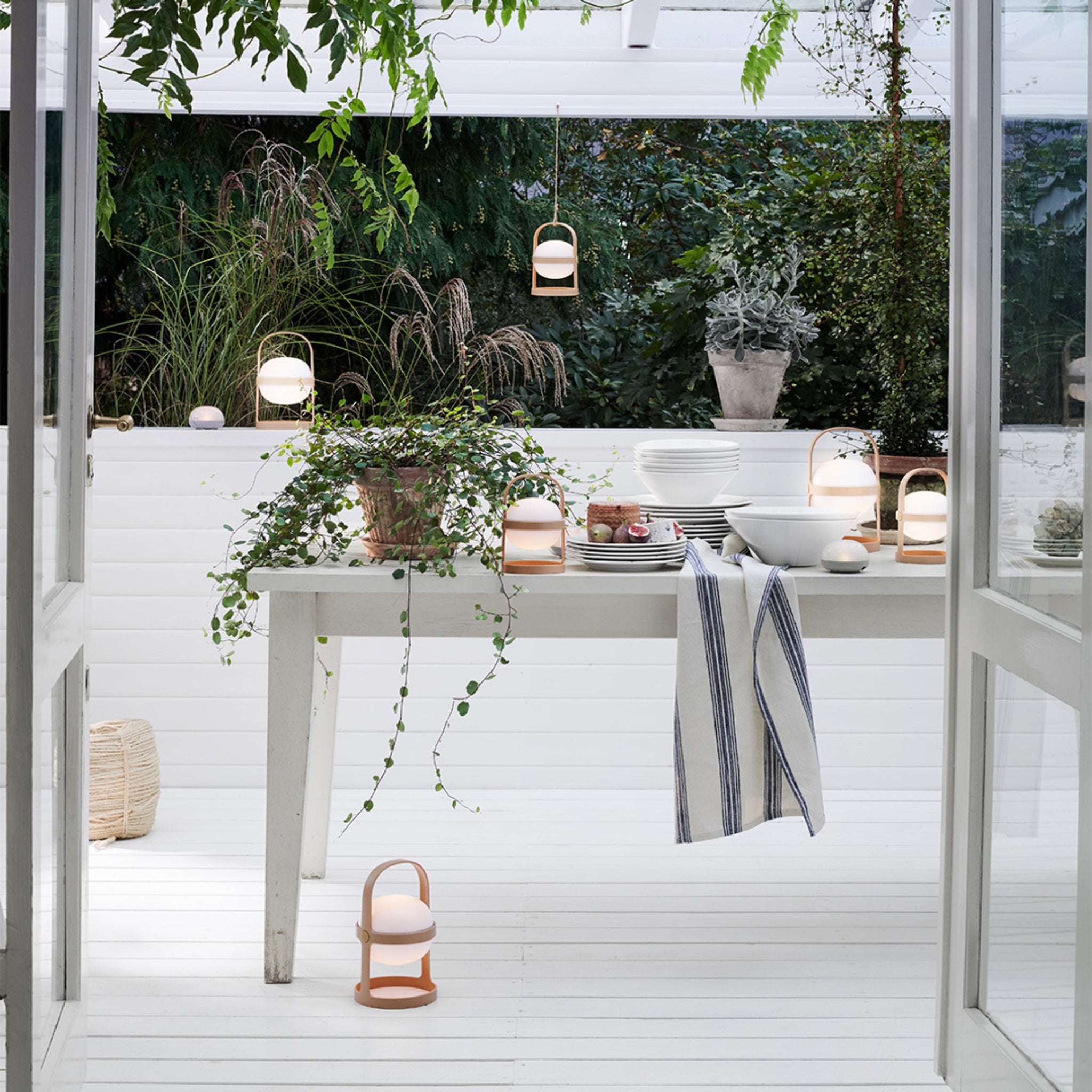Aan een witte eettafel buiten staan borden, schalen en planten, met lantaarns voor zacht licht. Groen gebladerte omringt de patio, waar een Rosendahl Garn Theedoek Blauw 50/70 over de rand is gedrapeerd, een toonbeeld van Scandinavische stijl.