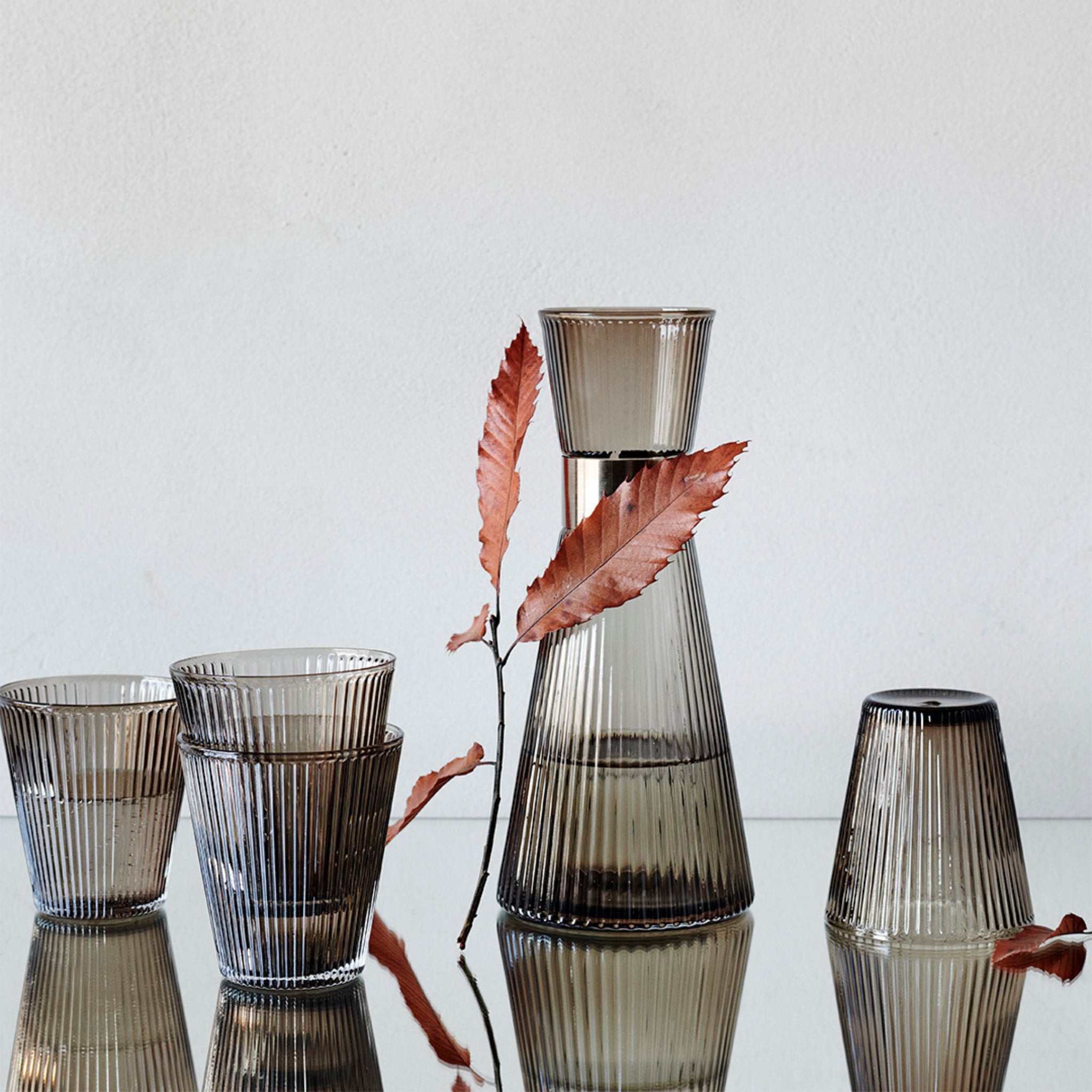 Een geribbelde, rokerige glazen set uit de Grand Cru-collectie van Rosendahl bevat de Grand Cru Karaf Walnoot 1 l en drie bekers, weergegeven met een roodbruin takje op een reflecterend oppervlak tegen een lichte achtergrond.