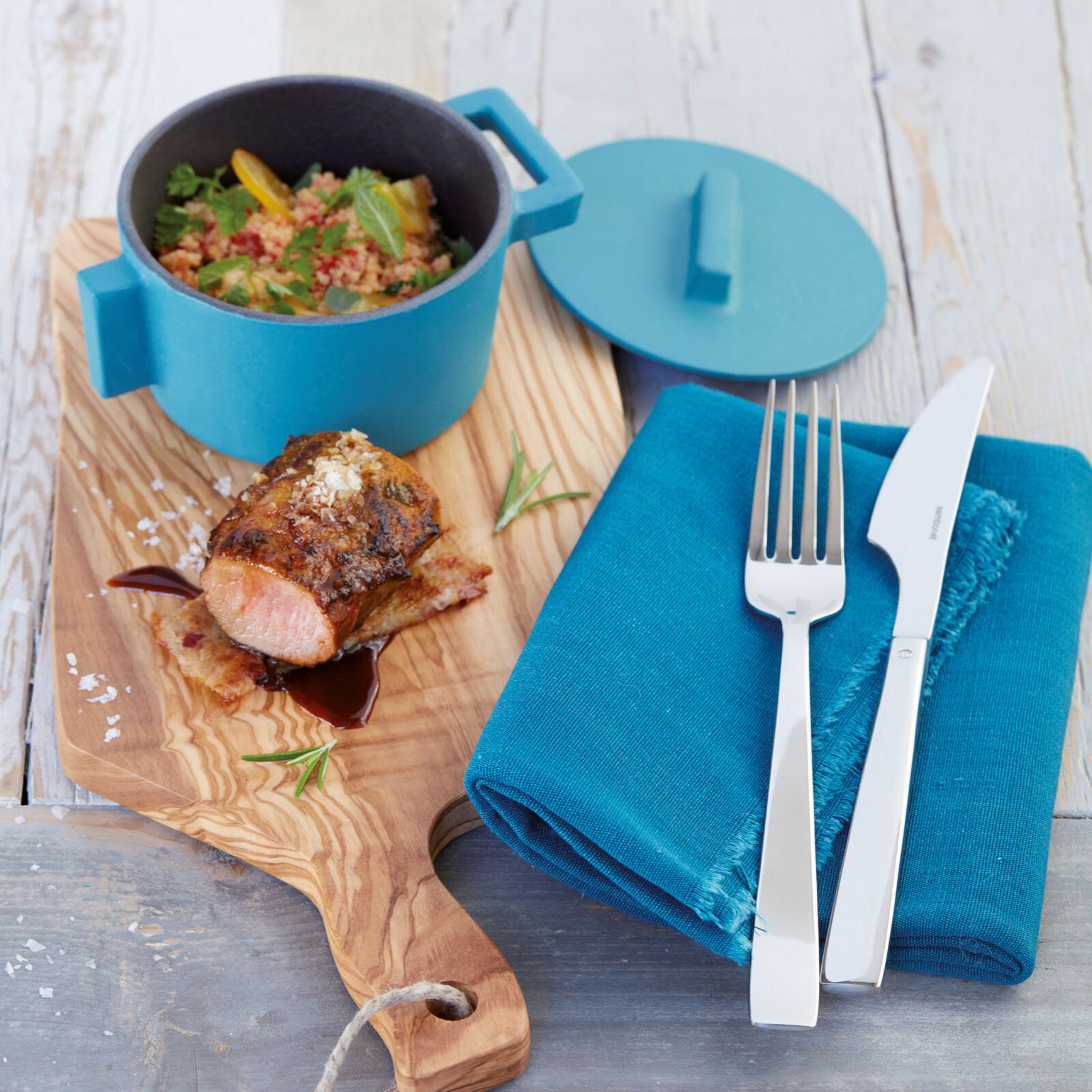 Op een houten plank staan een potje couscous met groenten, een plakje gekookt vlees met saus, een blauw servet en een bestekset Flat Bestek 24-Delig van Sambonet, allemaal op een licht houten oppervlak.