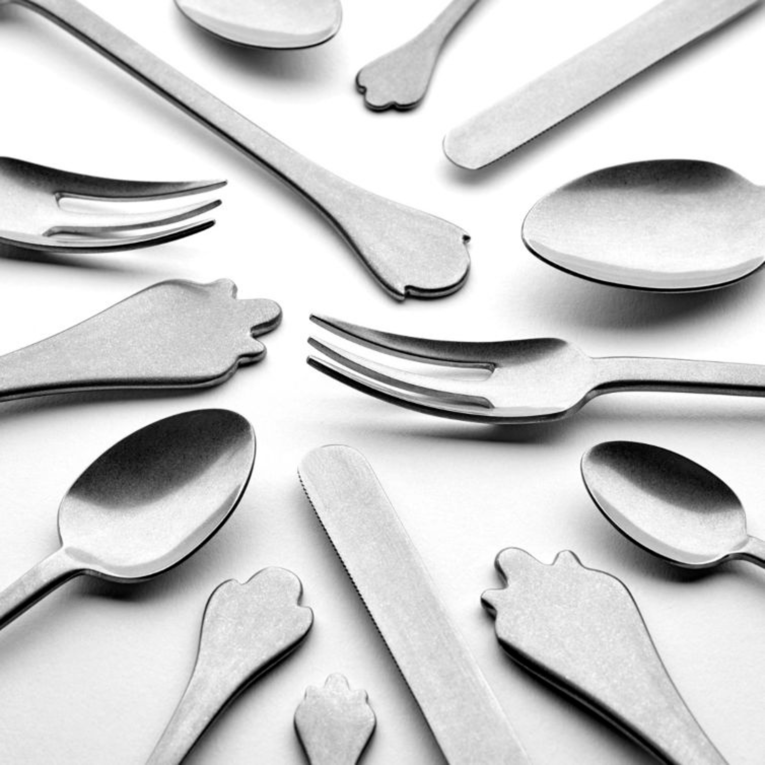Allerlei zilverwerk, waaronder de Serax Merci Bestek 24-delig set, is in een cirkel gerangschikt op een wit oppervlak, waarbij sommige gebruiksvoorwerpen aan de uiteinden zijn voorzien van speelse handgevormde ontwerpen.